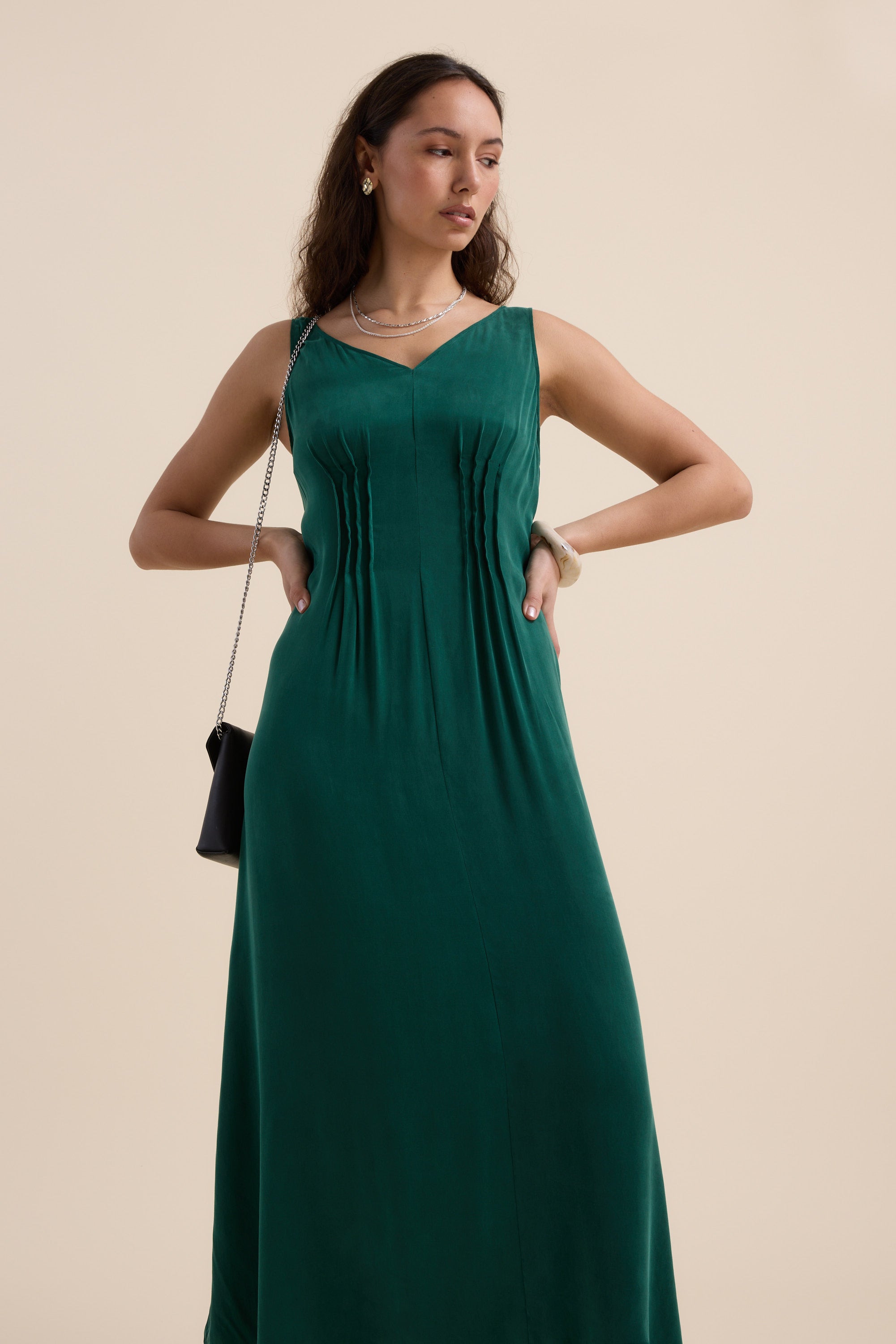 A woman wears the Max Cara Pleat Detail Dress, a sleeveless emerald green style with a cinched waist, paired with a black shoulder bag and jewelry, standing against a neutral beige background.
