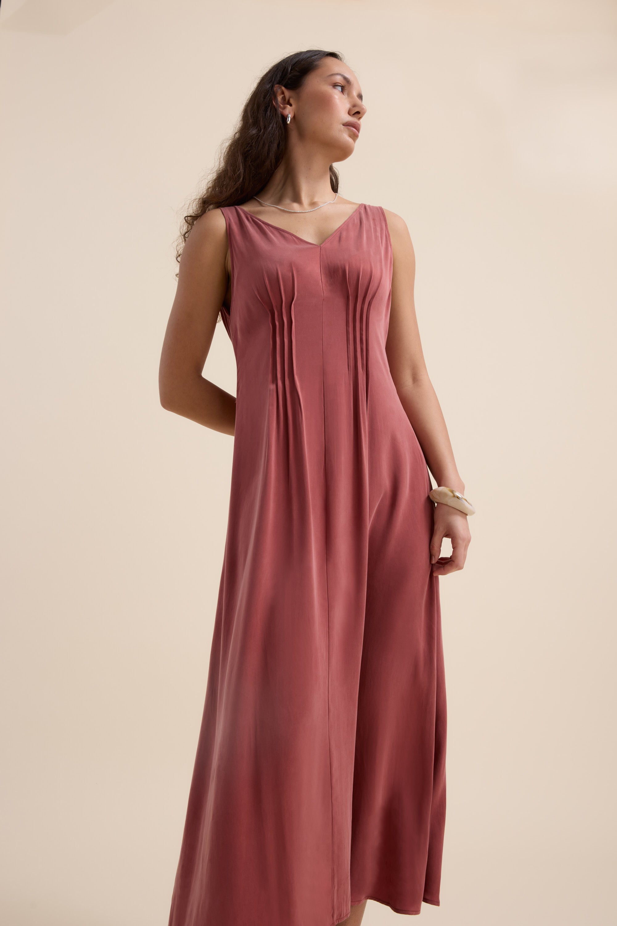Woman wears the Cara Pleat Detail Dress by Max in rose cupro blend, standing by a beige wall.
