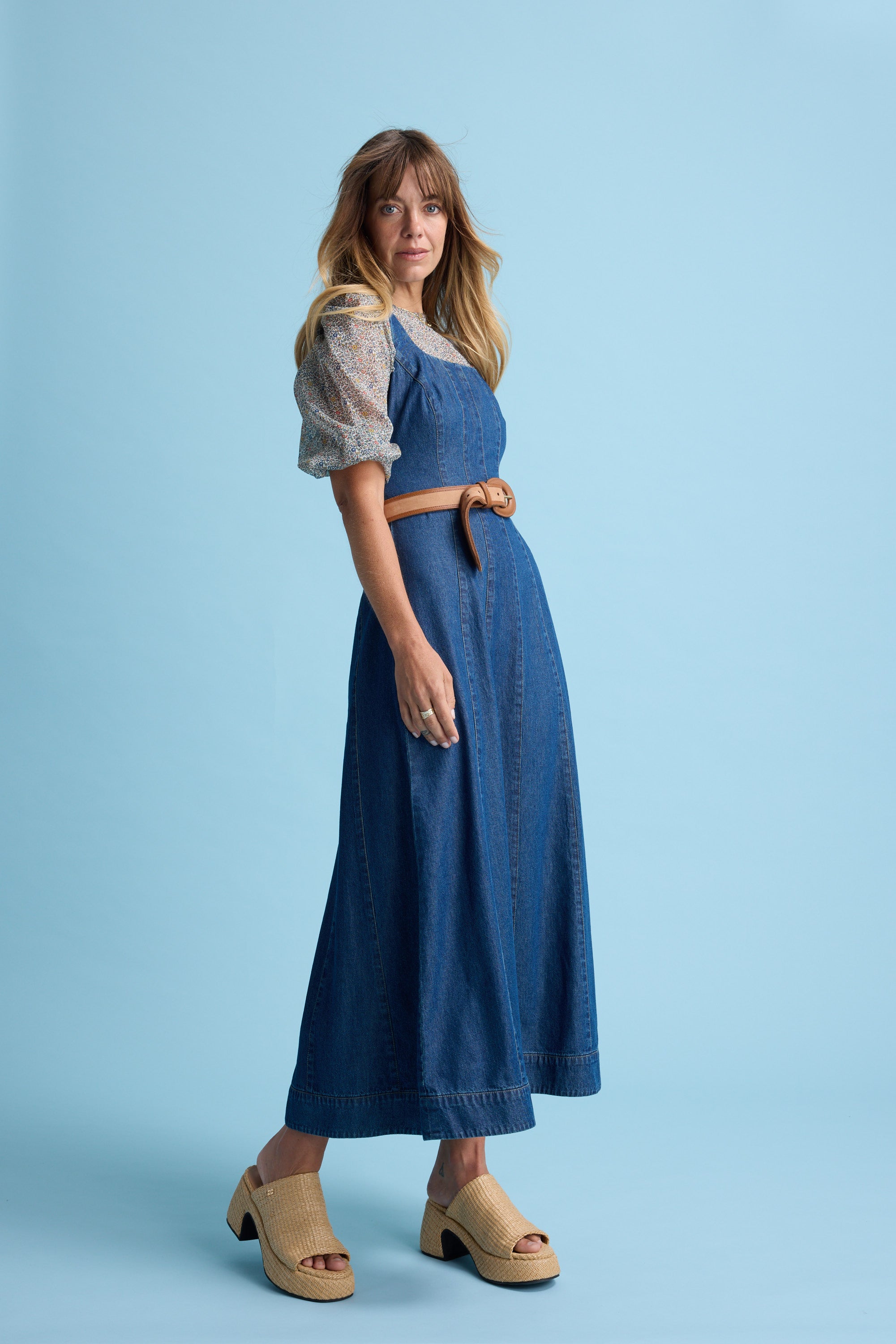 A woman with long, wavy blonde hair and a relaxed expression stands against a light blue background wearing the Eloise Puff Sleeve Blouse by Max, styled with a brown belt and beige platform sandals.