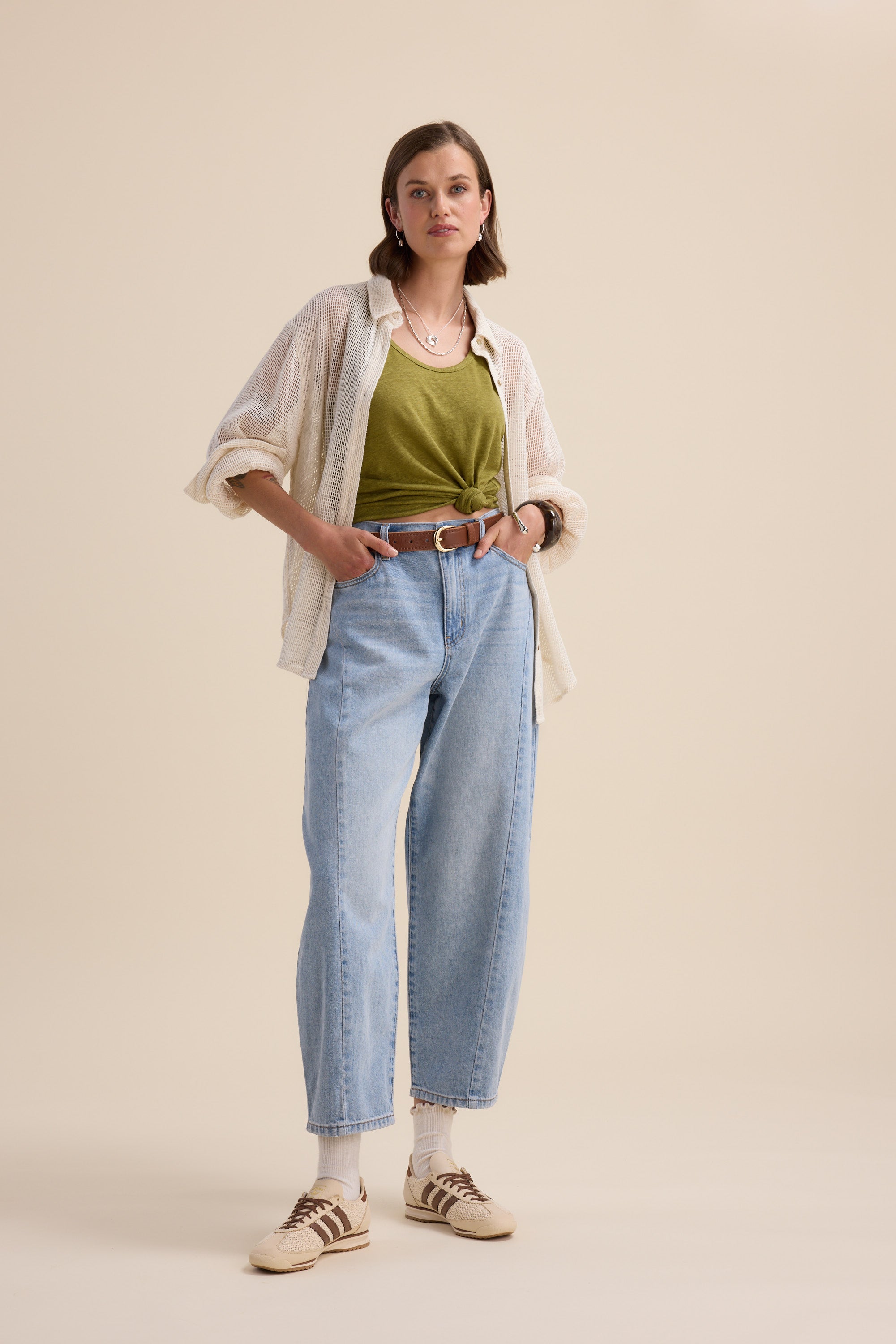 A person with short hair and a neutral expression wears the Elementary Linen Jersey Tank by Elementary beneath an open shirt, paired with high-waisted light blue jeans, a brown belt, white socks, and beige sneakers against a plain beige background.