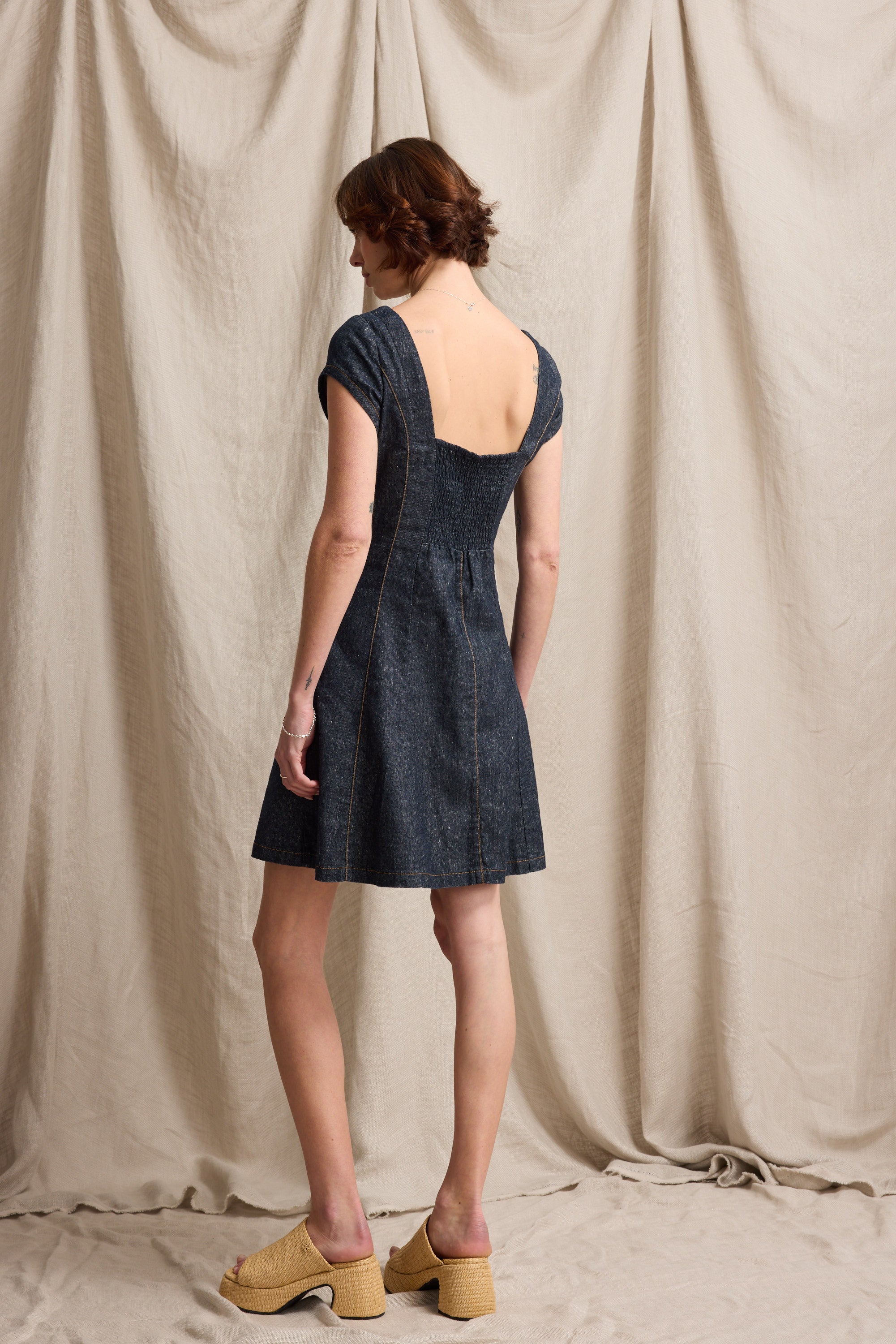 Woman wears Max's Linen Studio Cap Sleeve Dress, facing away against a beige backdrop.