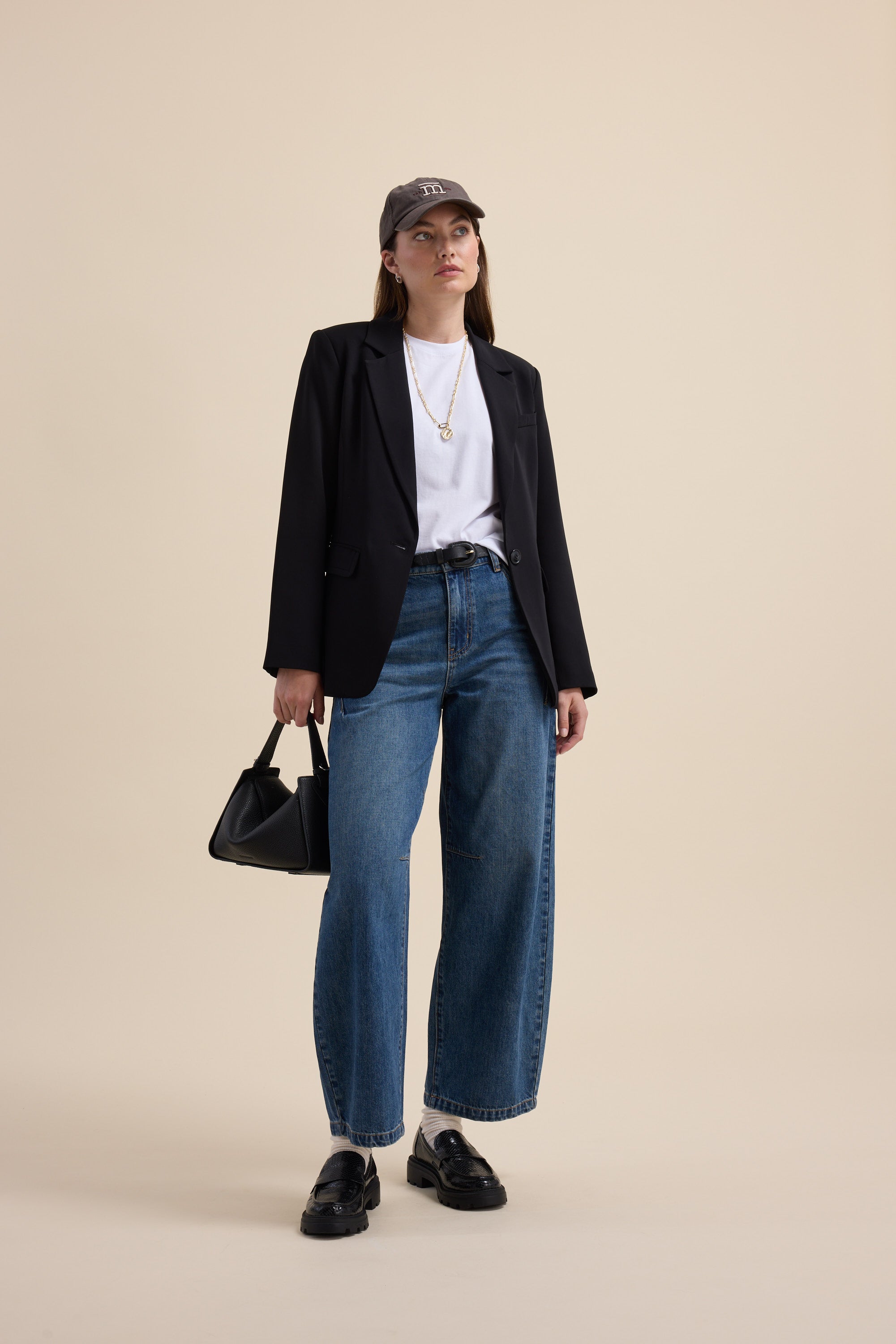 Woman in blazer, jeans, cap, and Max The Good Earth Cotton Crew Tee holds a black bag.