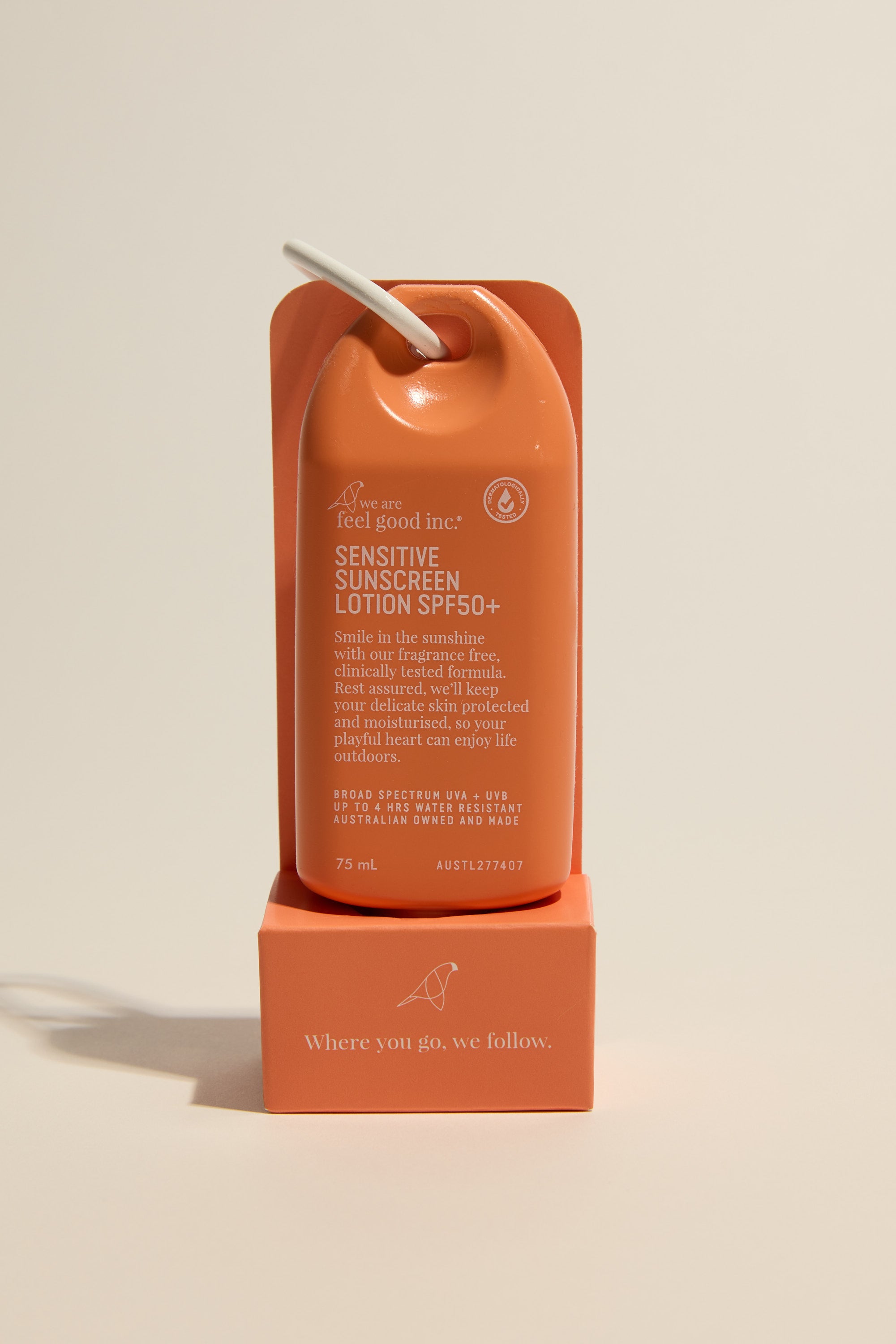 A bright orange bottle of We Are Feel Good Signature Sunscreen SPF50+ 75ml stands upright on a matching stand, with a white cap and loop. The broad spectrum sunscreen displays “Where you go, we follow.”.