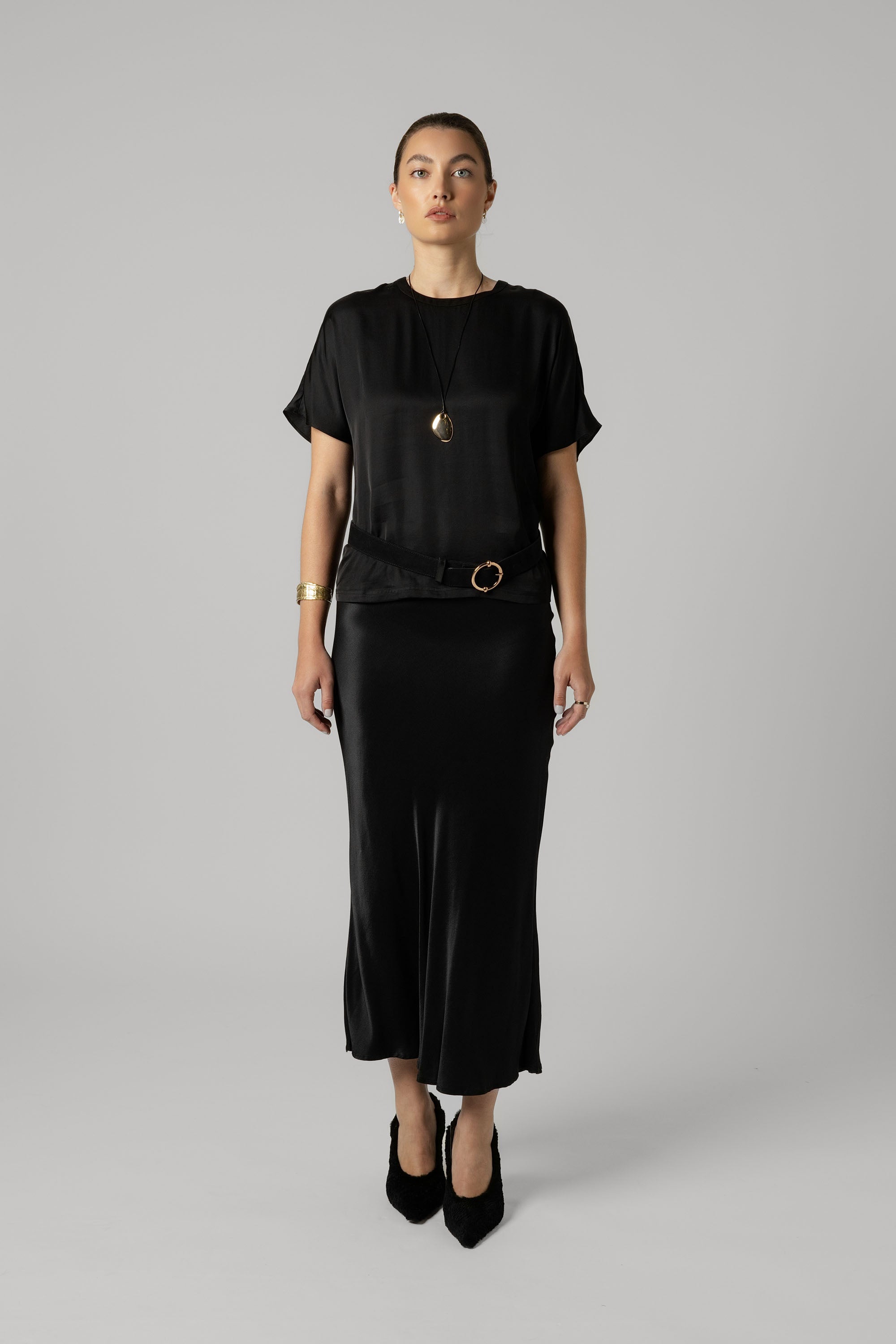 Woman in black top and Max's The Perfect Slip Skirt, with necklace and heels, on gray background.