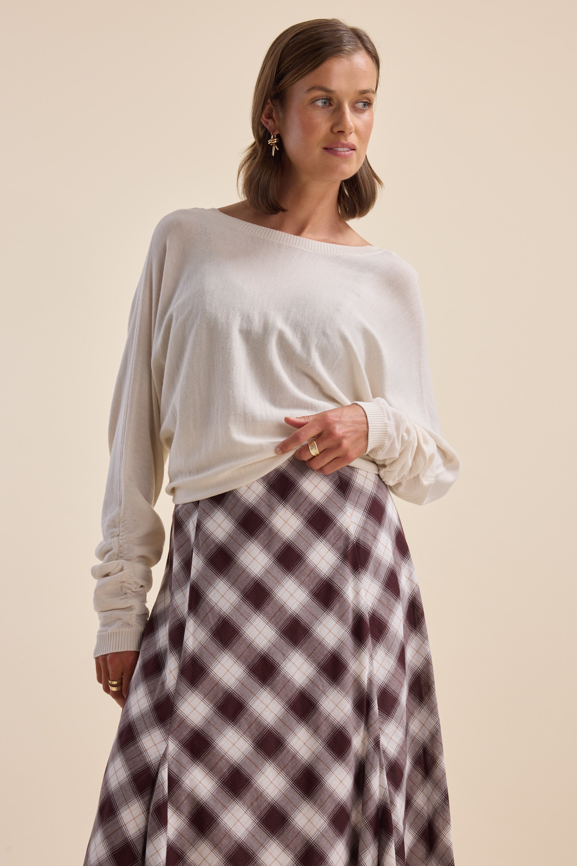 Woman in an Elementary ZQ Merino Reversible Sweater and plaid skirt against a beige background.