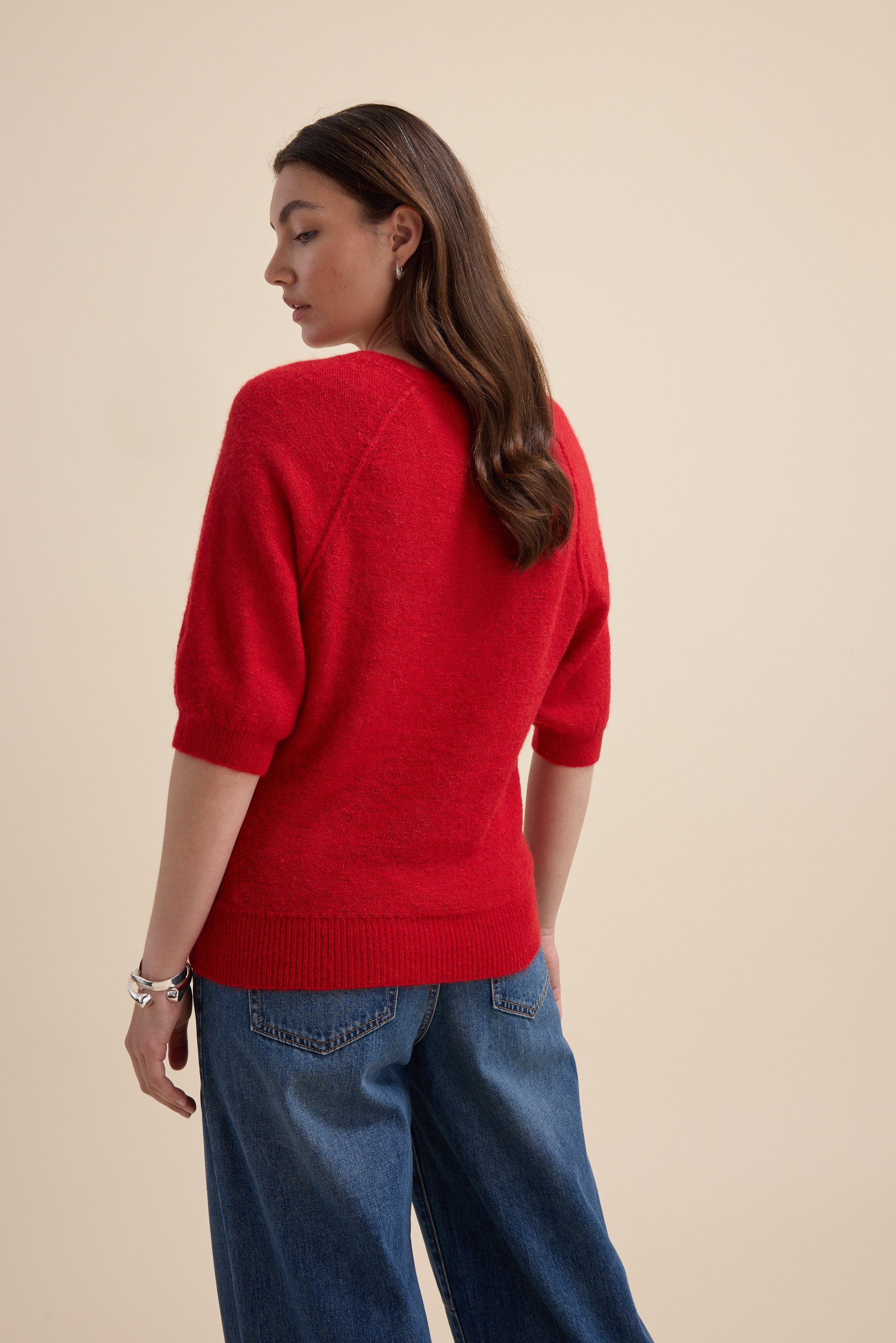 Woman wears a red Max Ivy Knitted V Neck Tee and blue jeans, back to the camera.
