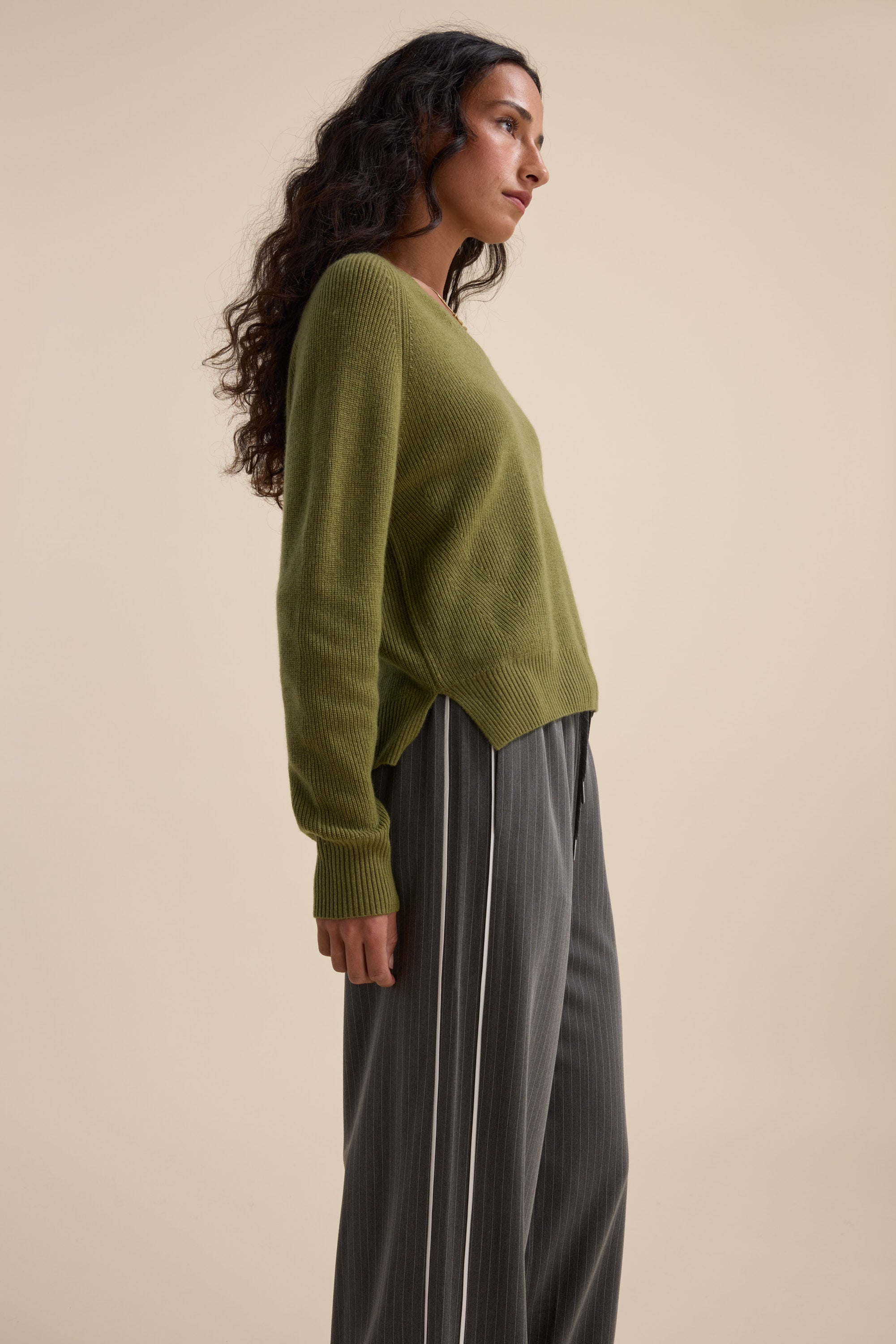 Person wears Elementary Cashmere Blend V Neck Sweater and gray pinstripe pants on beige background.