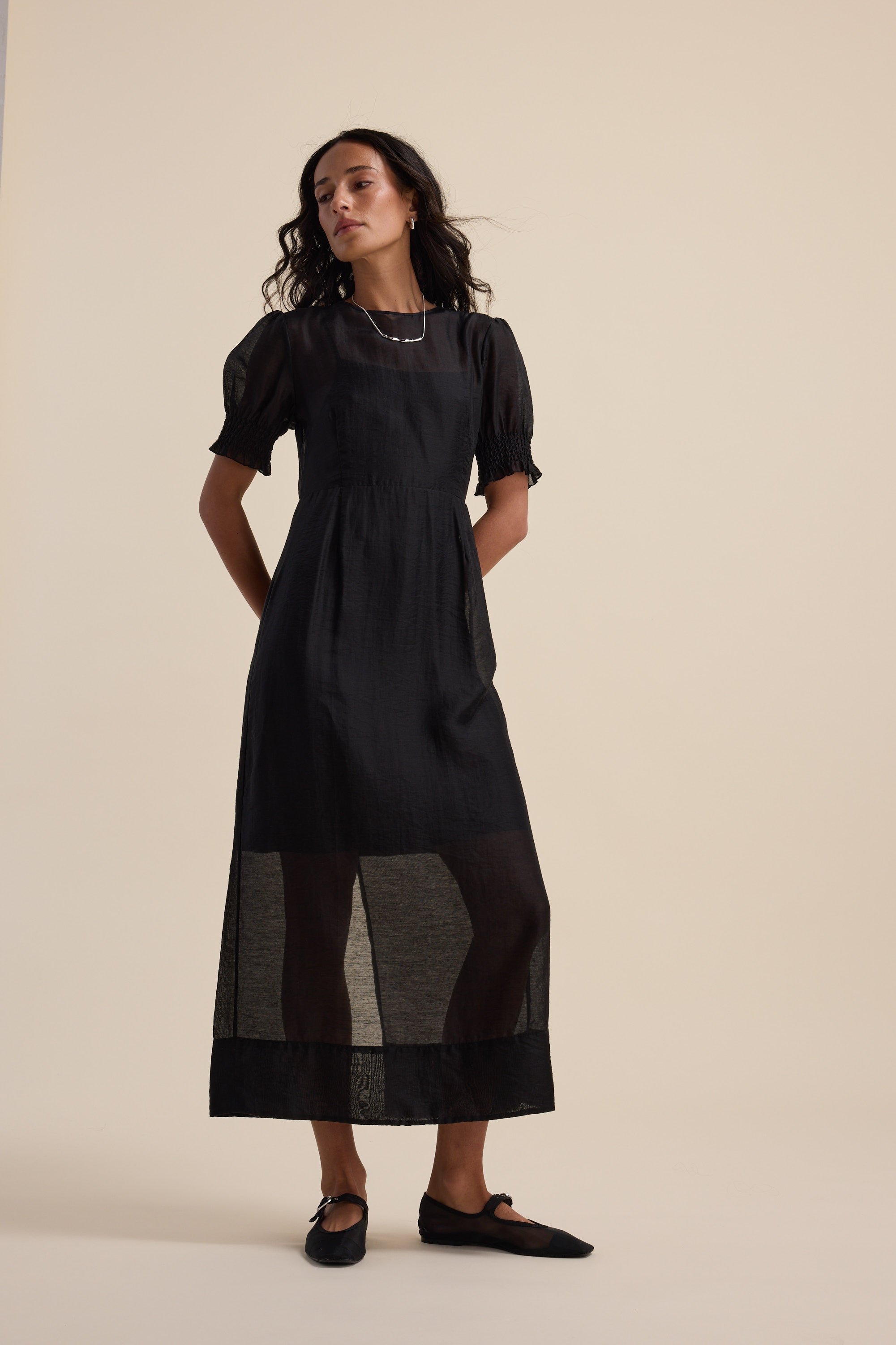 Model in Max’s The Sheer Layering Dress with black flats stands against a beige background.