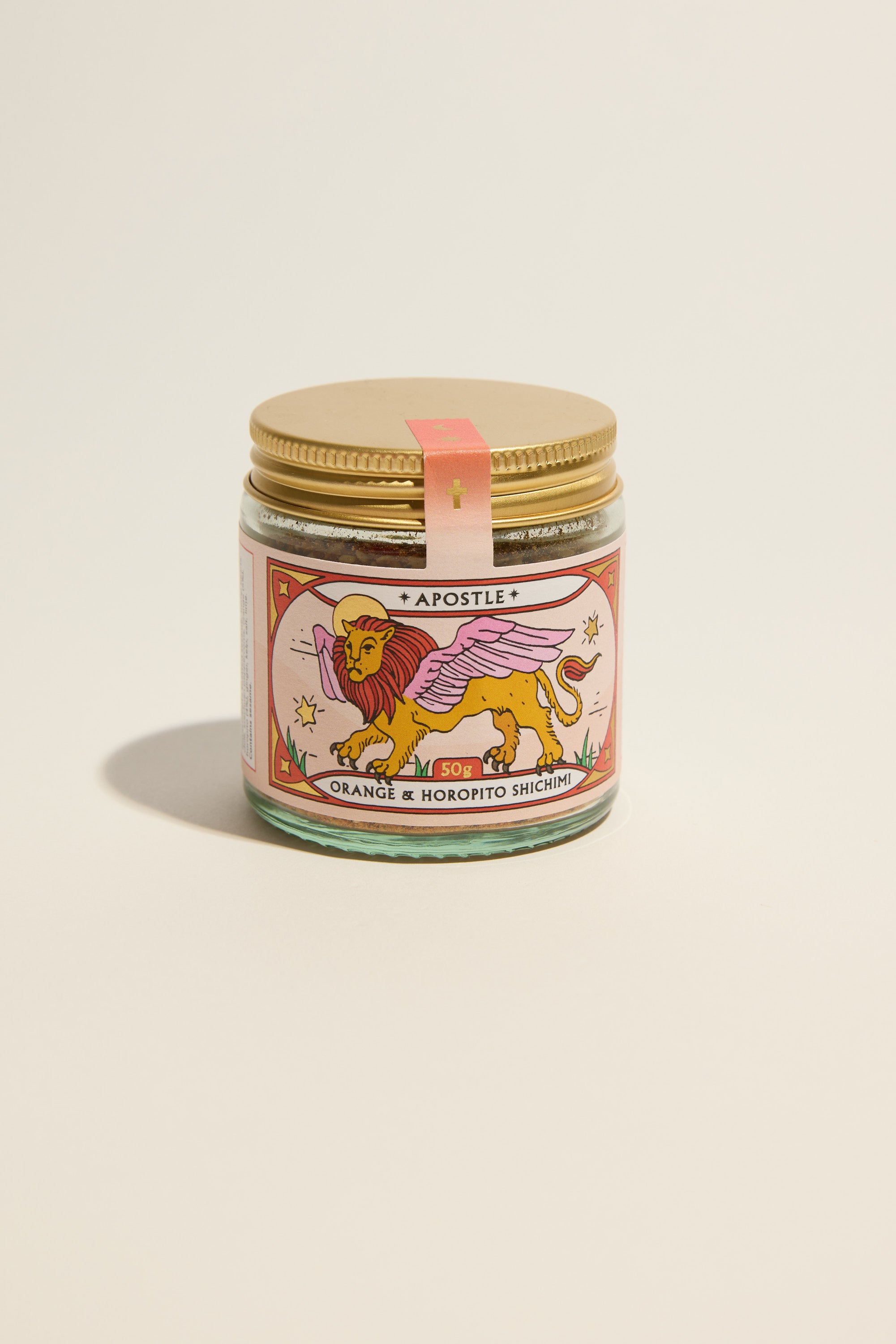 A glass jar with a gold lid and a pink label featuring a winged lion illustration sits on a neutral background. The label reads "Apostle Orange & Horopito Shichimi" by Apostle, inspired by Japanese Shichimi Togarashi spice blend.