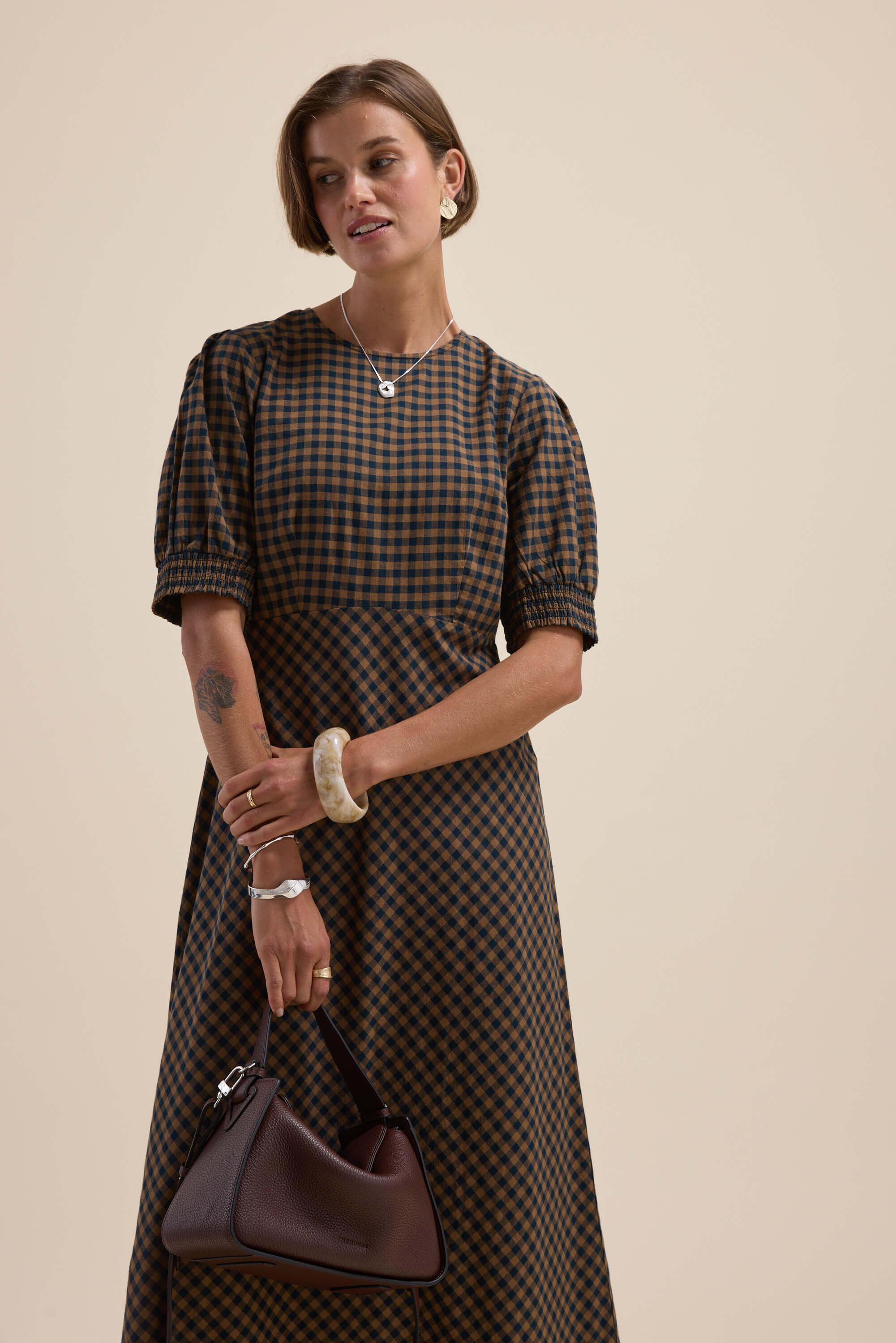 Woman in Max Lucy Bias Cut Dress holds dark handbag, wears jewelry, beige background.