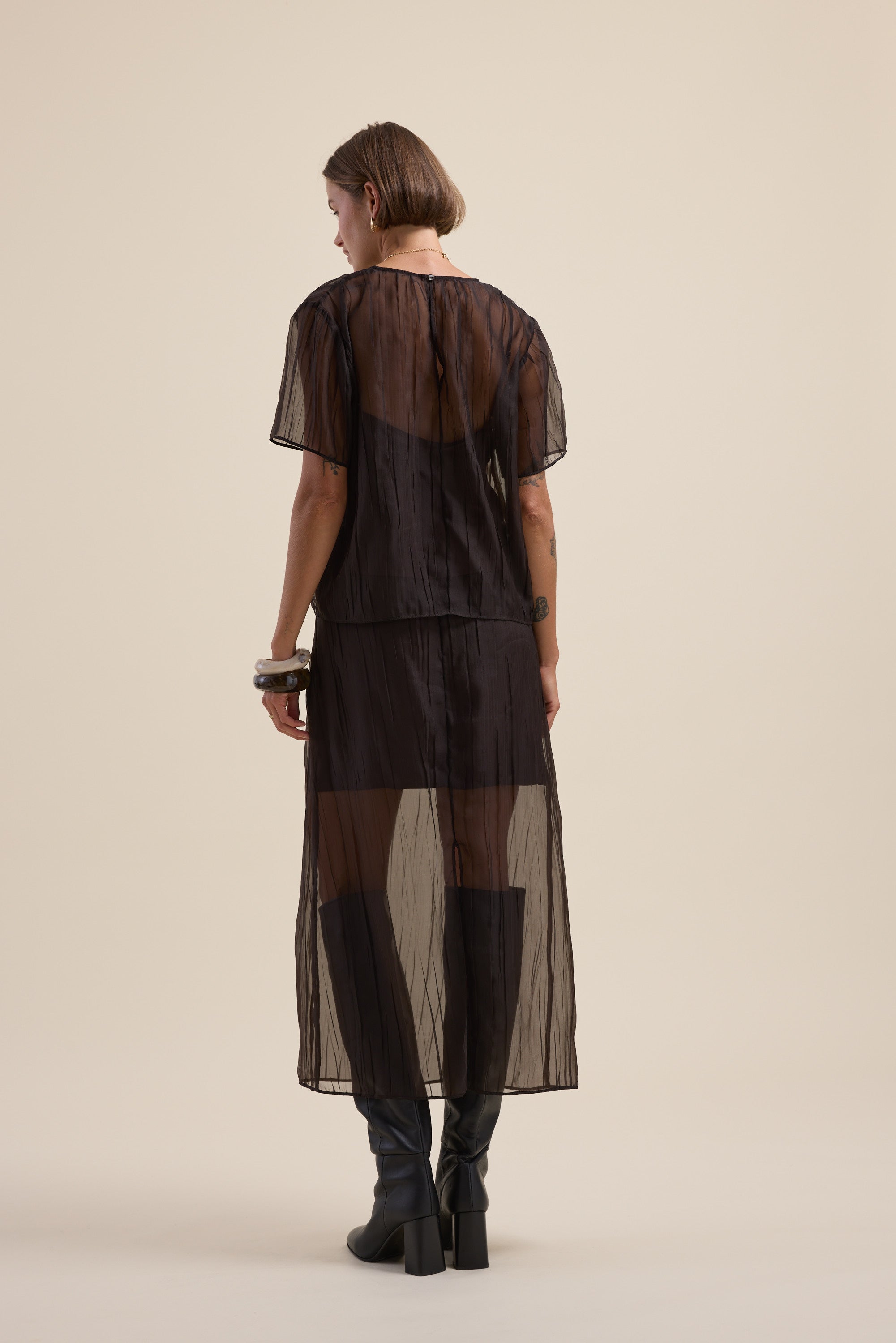 Person wears Max Ari Sheer Midi Skirt and boots, back to camera, beige background.