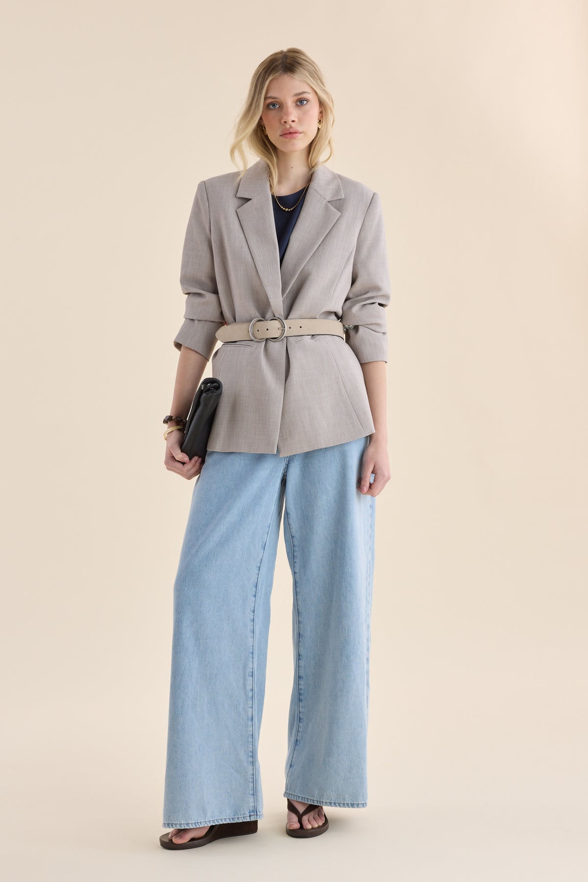 Woman in Max’s The Relaxed Blazer, wide jeans, black sandals, with clutch on beige background.