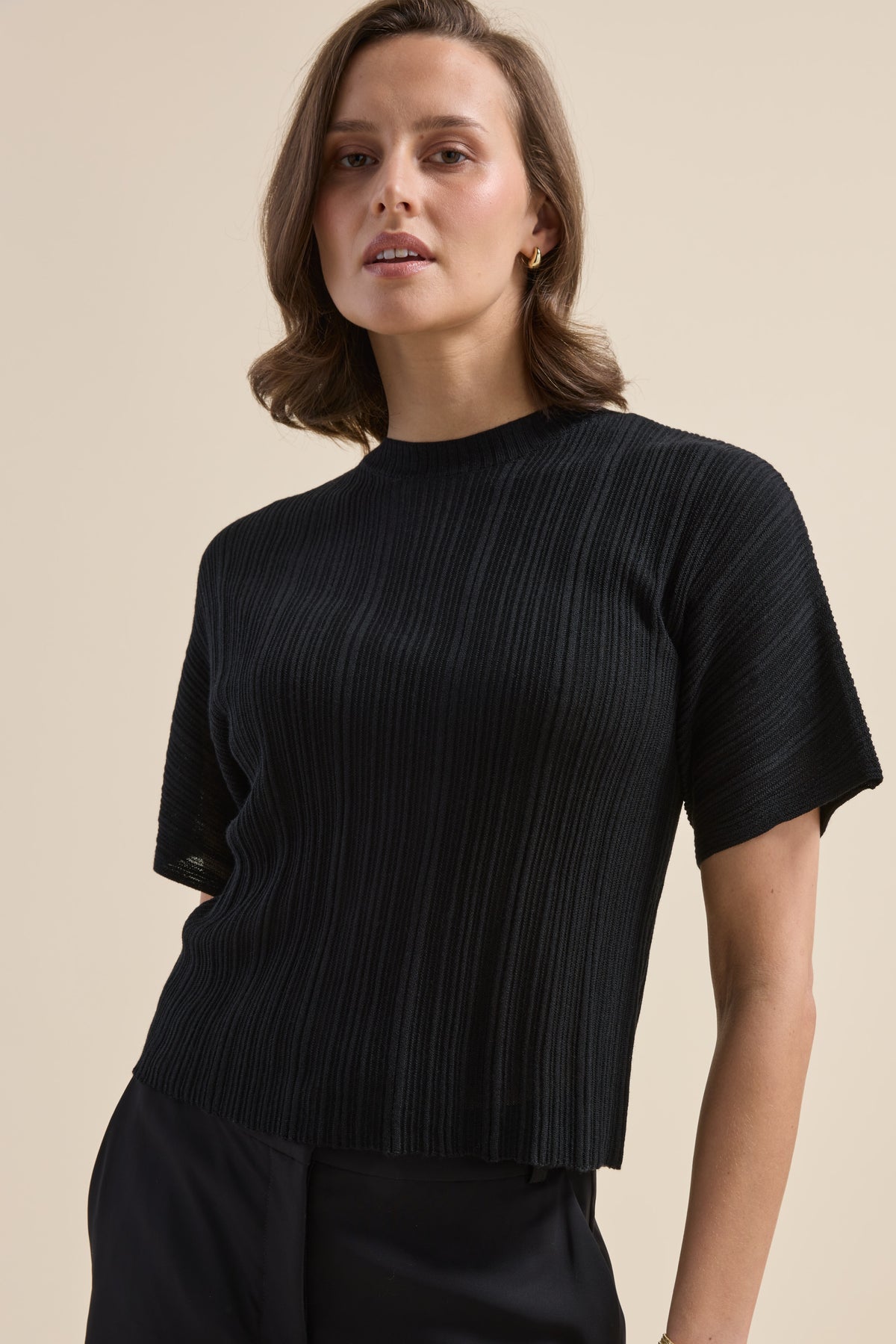 Woman in Max’s Merino Silk Knit Detail Tee, short-sleeved, black, with dark pants on beige bg.