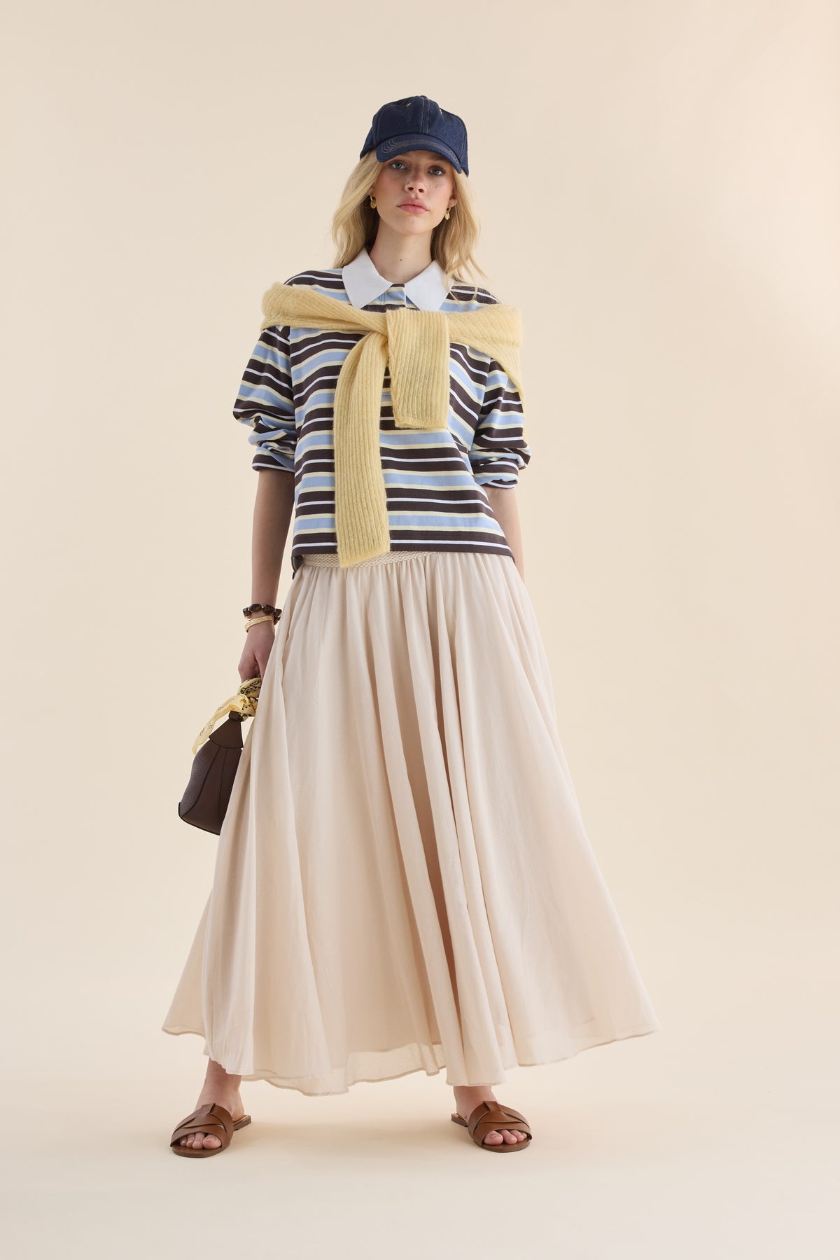 Woman in Elementary Ice Blue Stripe Polo, cream skirt, blue cap, sandals, brown bag.