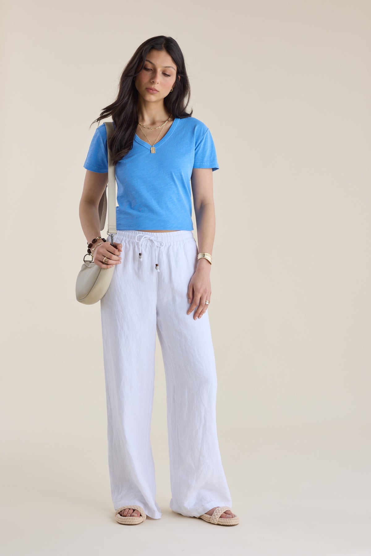 Woman in Elementary Organic Cotton Slub V Neck Tee, white pants, sandals, bag, beige backdrop.