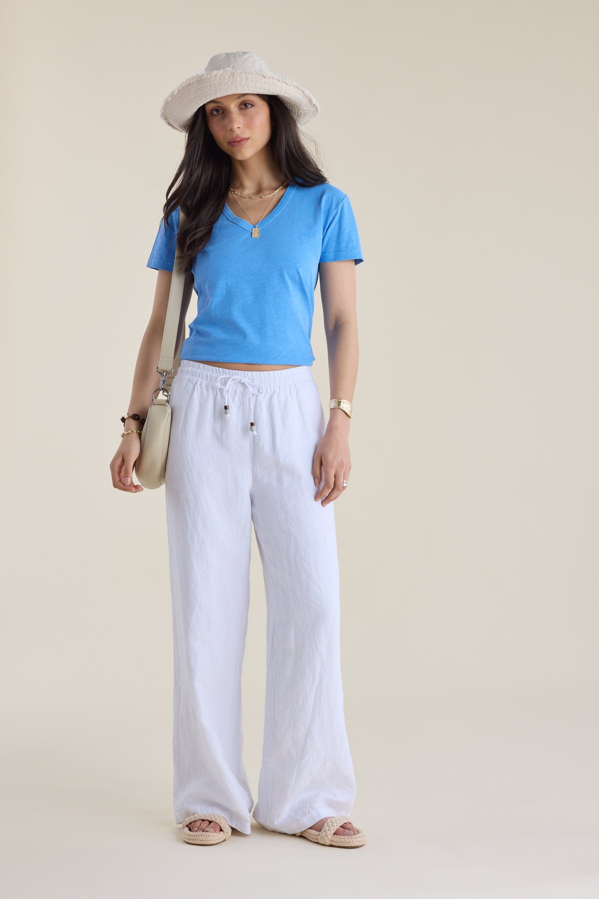 Woman in Elementary Organic Cotton Slub V Neck Tee, white pants, sunhat, sandals, plain backdrop.