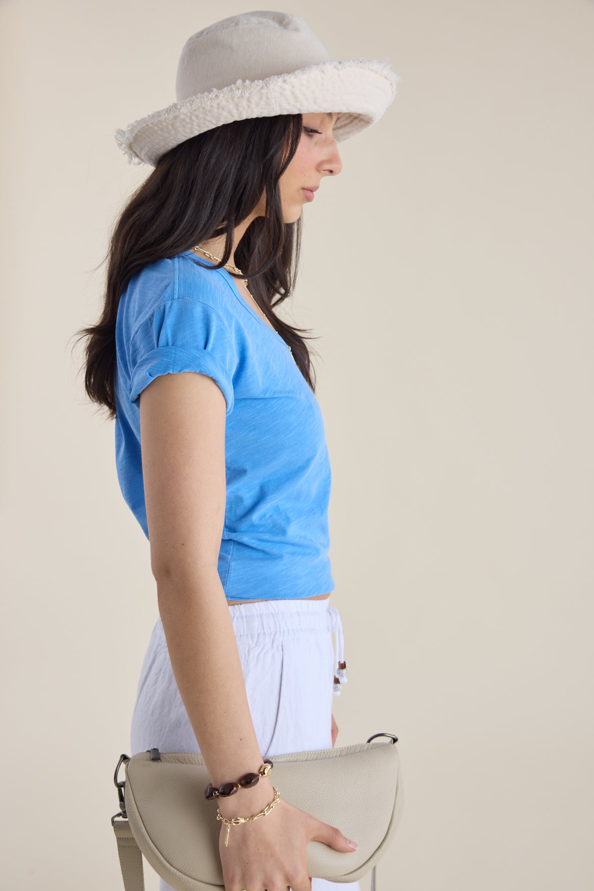 Woman in Elementary Organic Cotton Slub V Neck Tee, hat, and beige purse, plain background.