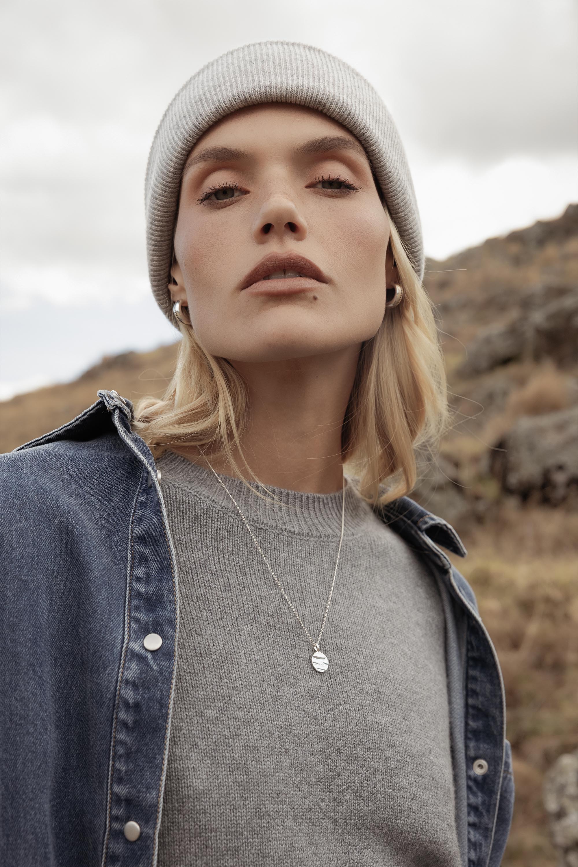 A person with blonde hair stands outdoors on rocky terrain, wearing a light gray beanie, gray sweater, denim jacket, small earrings, and the Caja Isla Circle Necklace from Caja in recycled sterling silver, looking confidently at the camera.