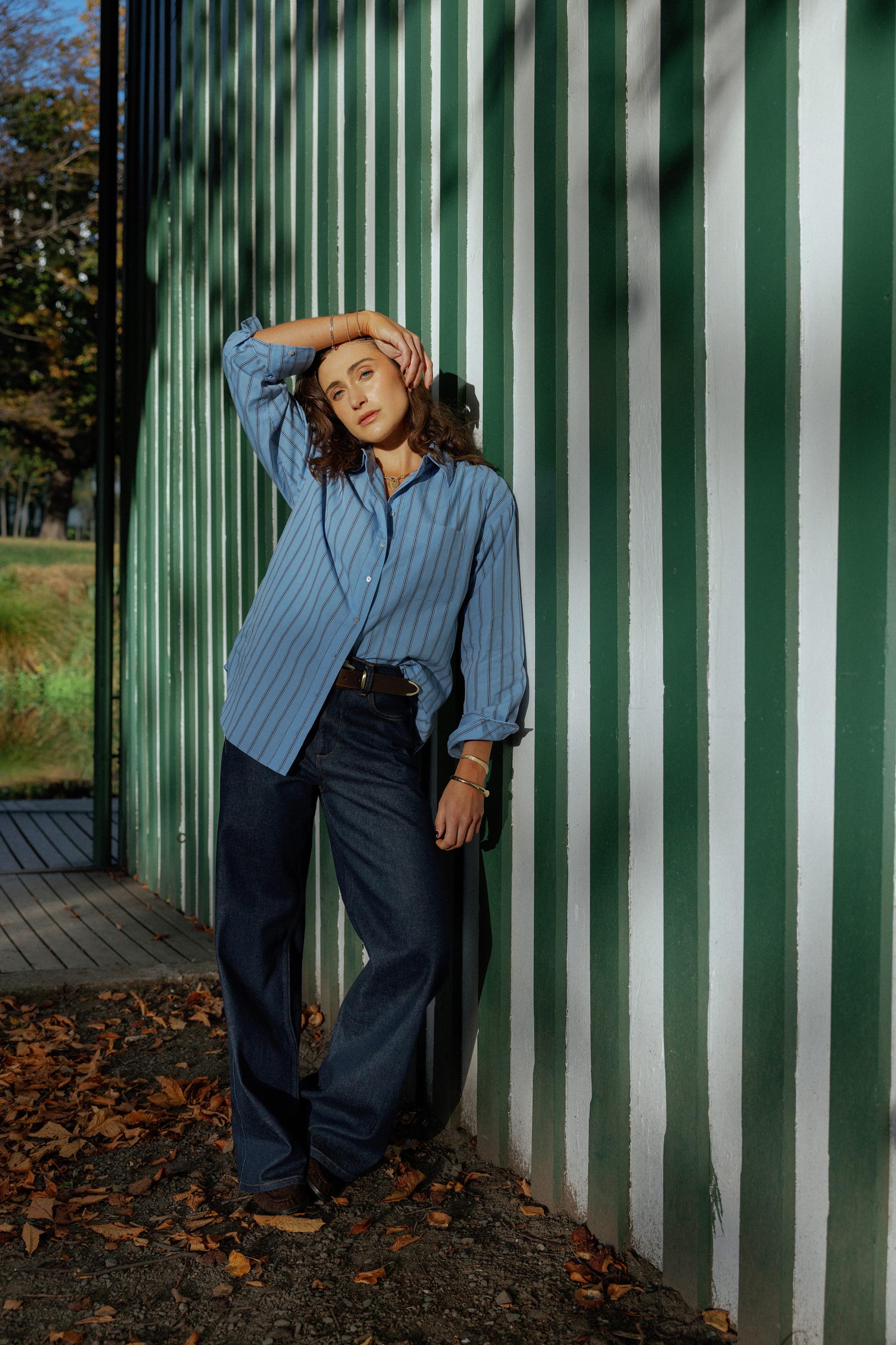 Woman in Nevada Wide Jeans by Max leans on striped wall with autumn leaves nearby.