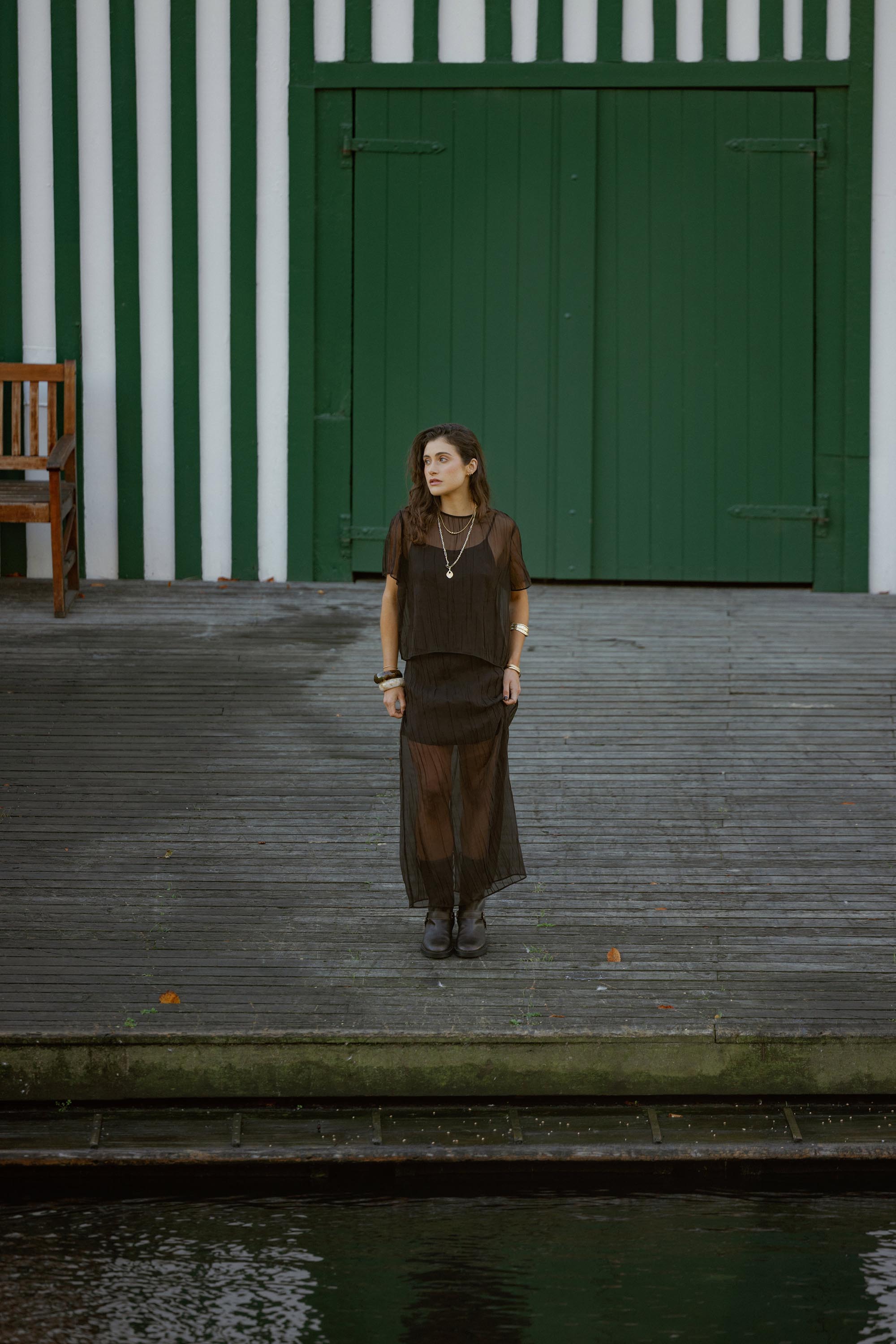 Woman in the Max Ari Sheer Midi Skirt stands on a wooden dock with green doors behind her.