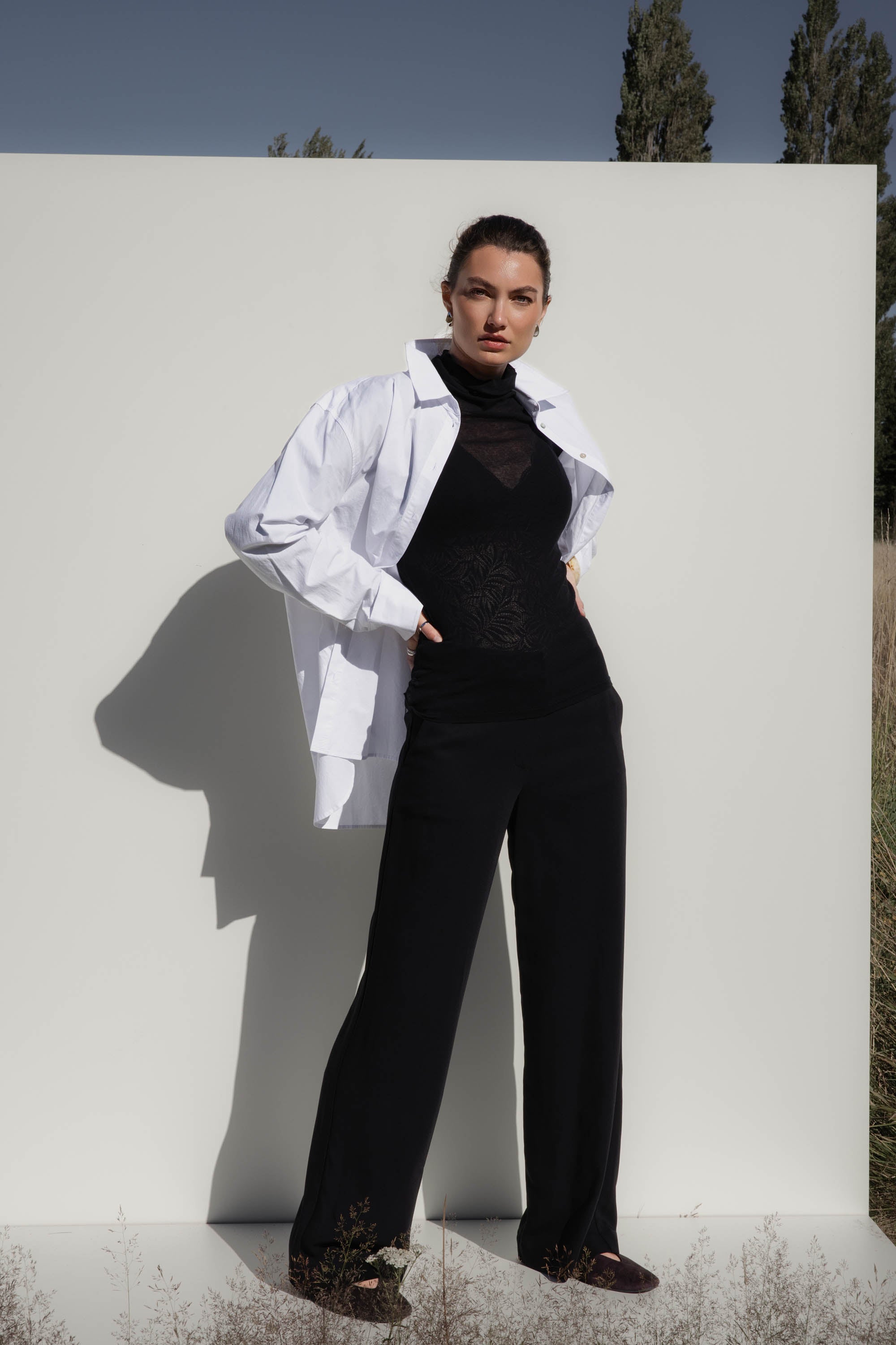 Woman poses outdoors in Max's The Ultimate Wide Leg Pants with a white shirt over her shoulders.