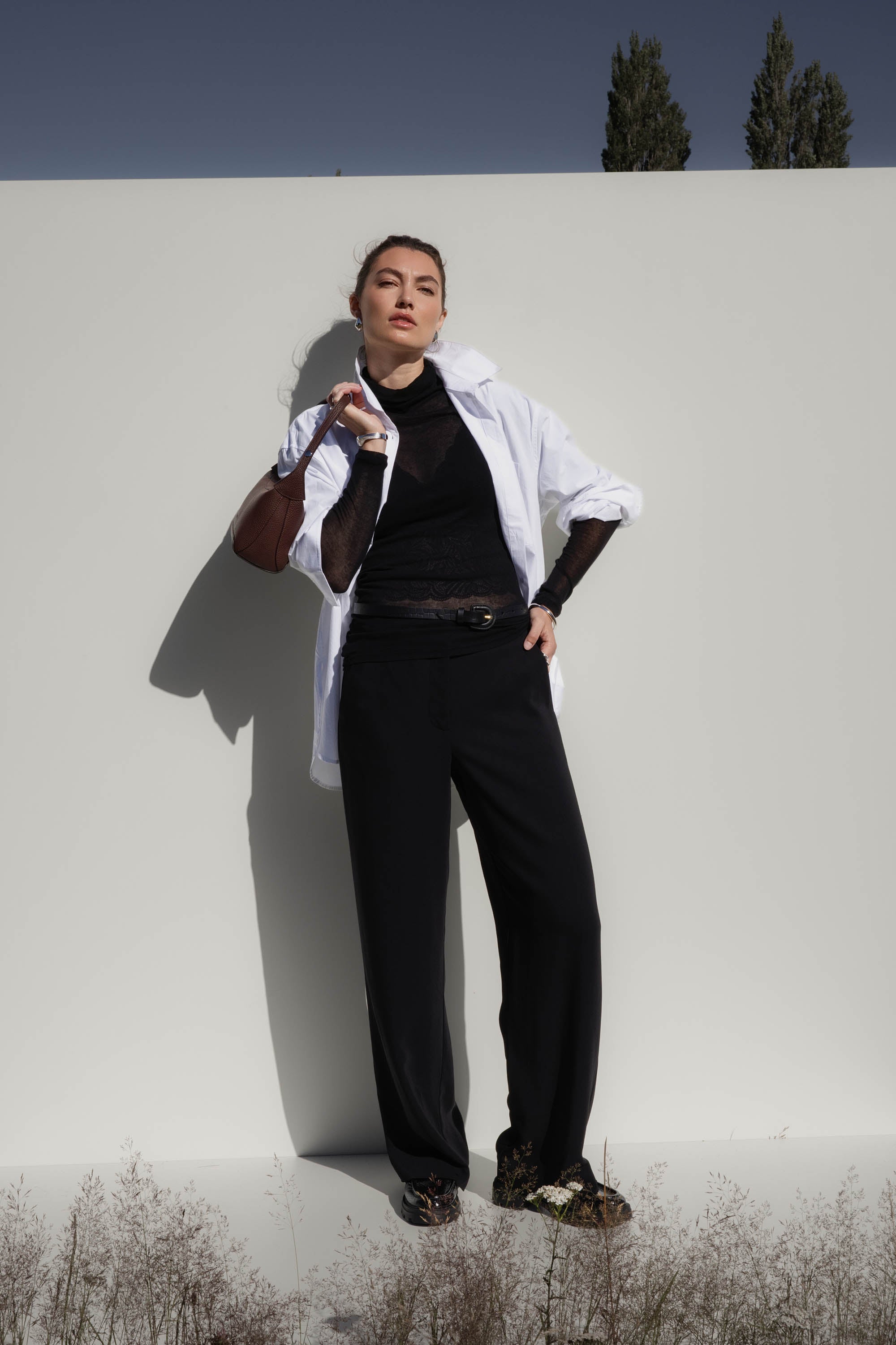 Woman wears Max's The Ultimate Wide Leg Pants and white shirt, posing by a plain light wall.