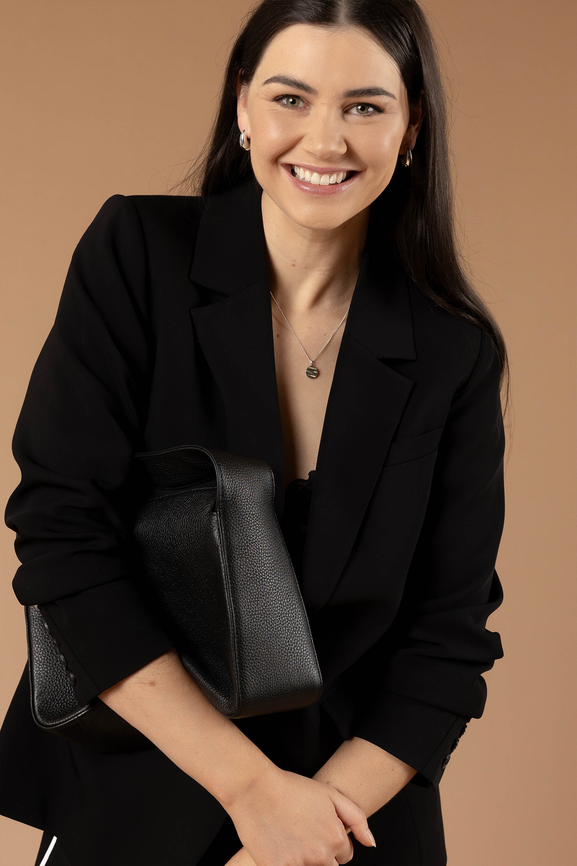 A woman with long dark hair smiles at the camera against a beige background, wearing Max’s The All Occasion Blazer and holding a black textured handbag.