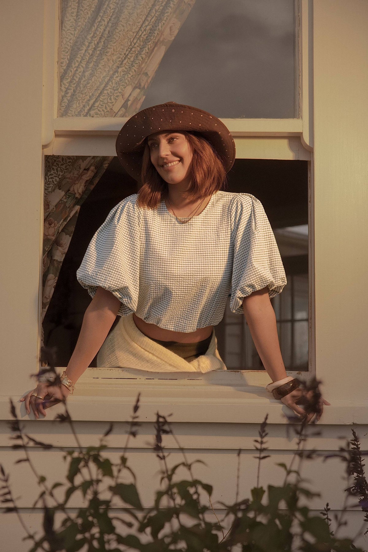 Woman in a hat and Max Coco Bubble Sleeve Top smiles while leaning out a window at dusk.