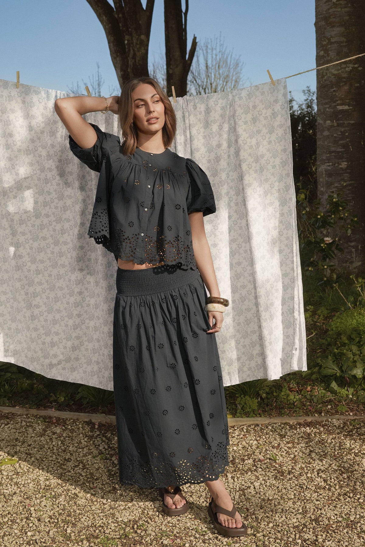 Woman in Elementary’s Broderie Detail Skirt stands outdoors by a laundry line with white sheets.
