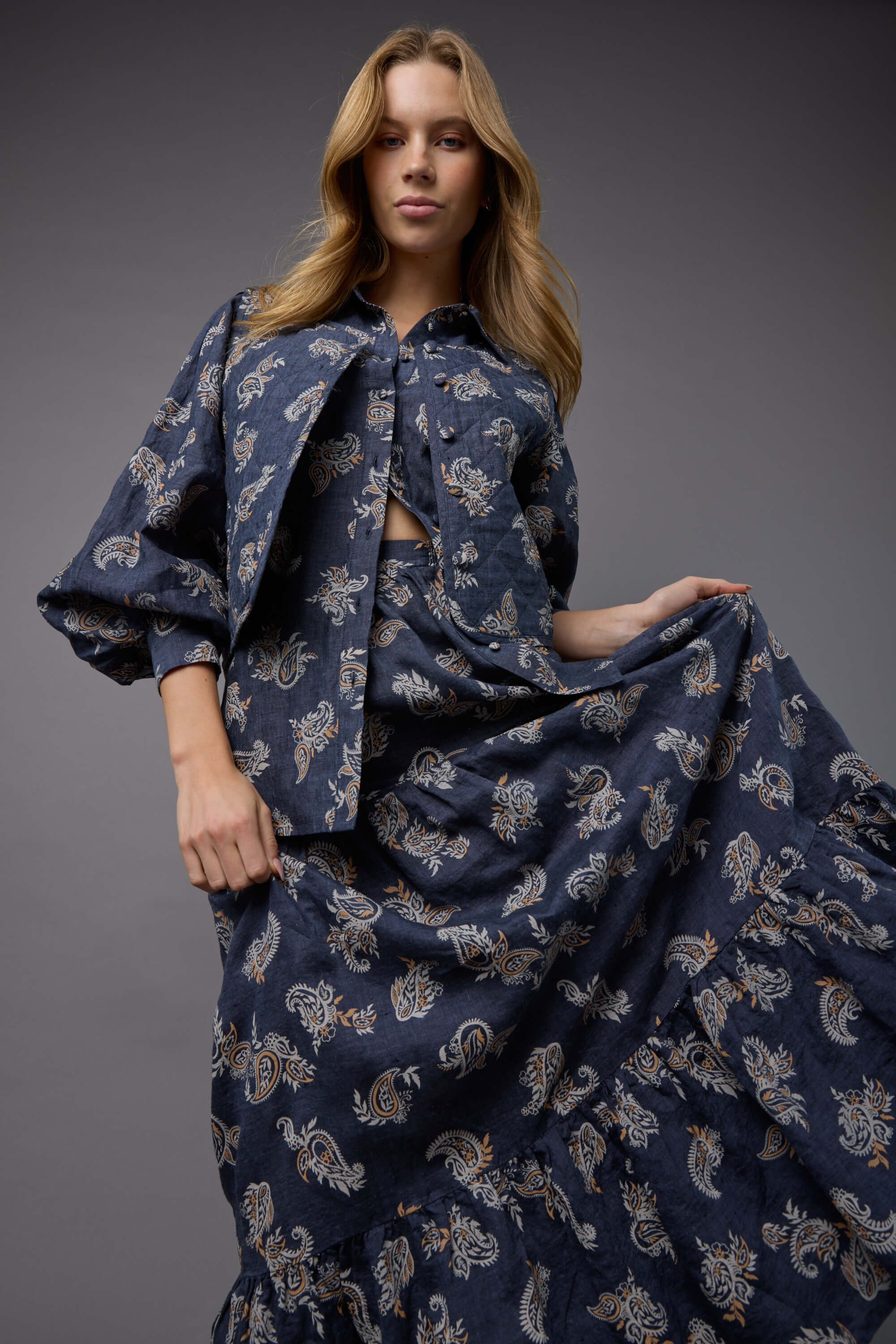 A woman models the Max Quinn Tiered Skirt and a matching dark blue button-up shirt with paisley print, holding out the pleated maxi skirt against a neutral gray backdrop.