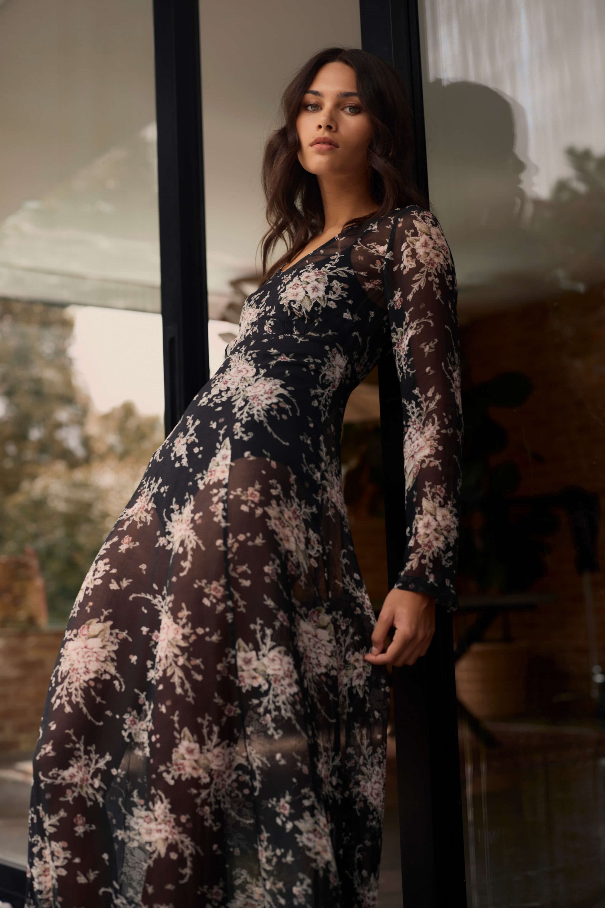 A woman with long, wavy brown hair stands by a glass door wearing the Amber Long Sleeve Dress by Max, featuring a sheer fabric and light floral pattern. She gazes confidently at the camera, with daylight and greenery outside.