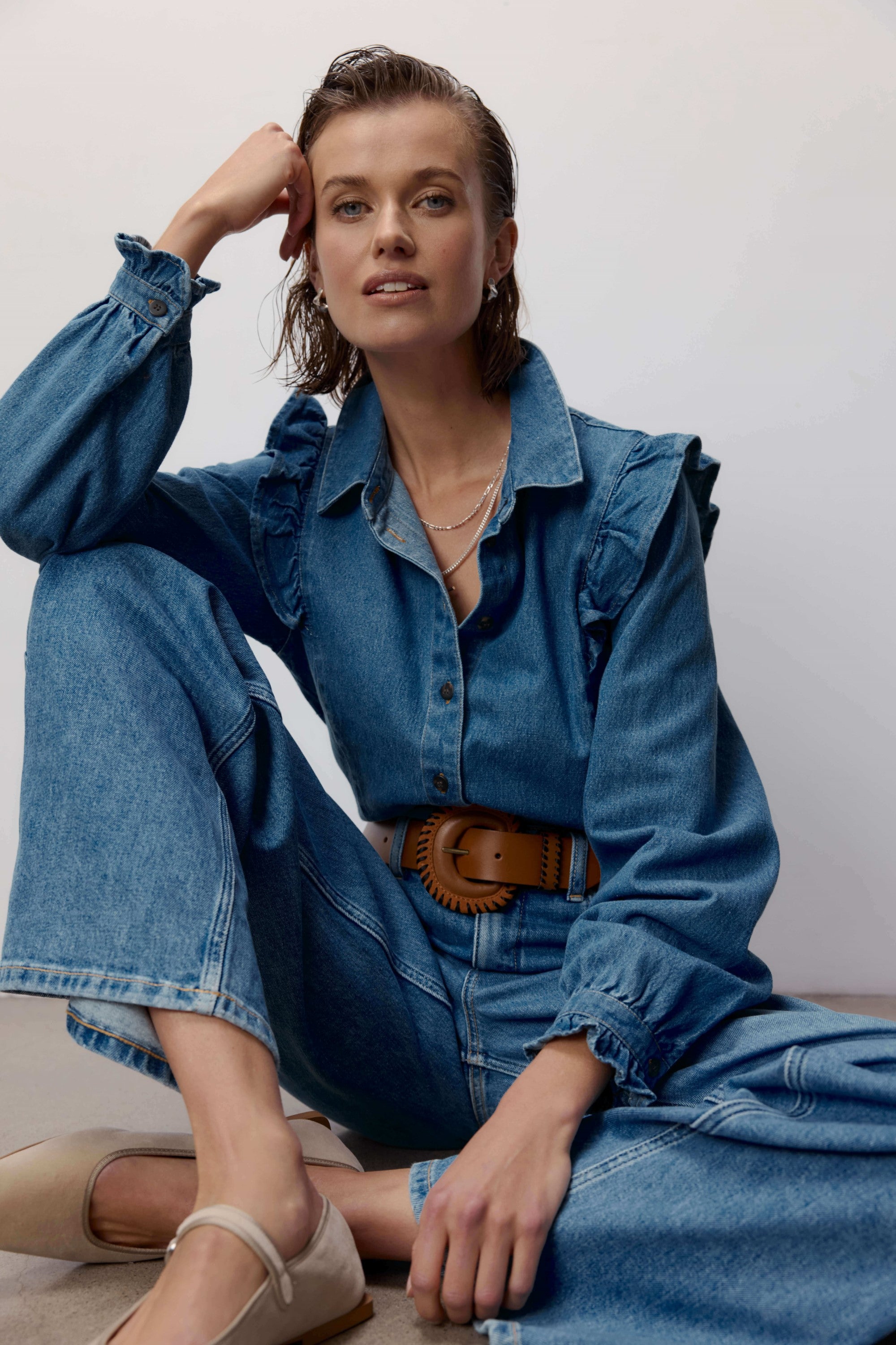A woman with wet, slicked-back hair sits on the floor wearing Max’s Nevada Wide Jeans and a blue denim shirt with ruffled details. She accessorizes with a brown belt, layered necklaces, and beige flats.