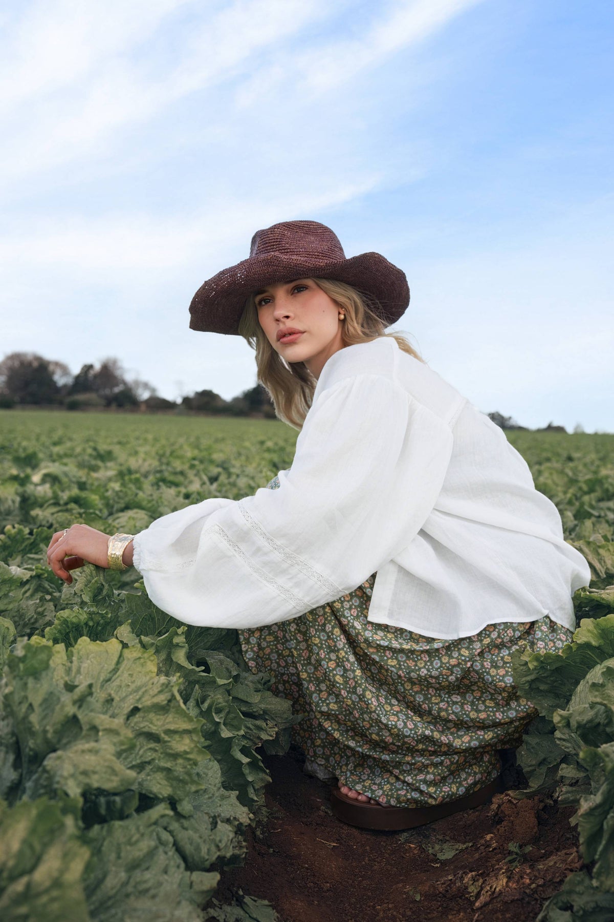 Woman in Millie Trim Detail Blouse by Max squats in a green field under a blue sky.