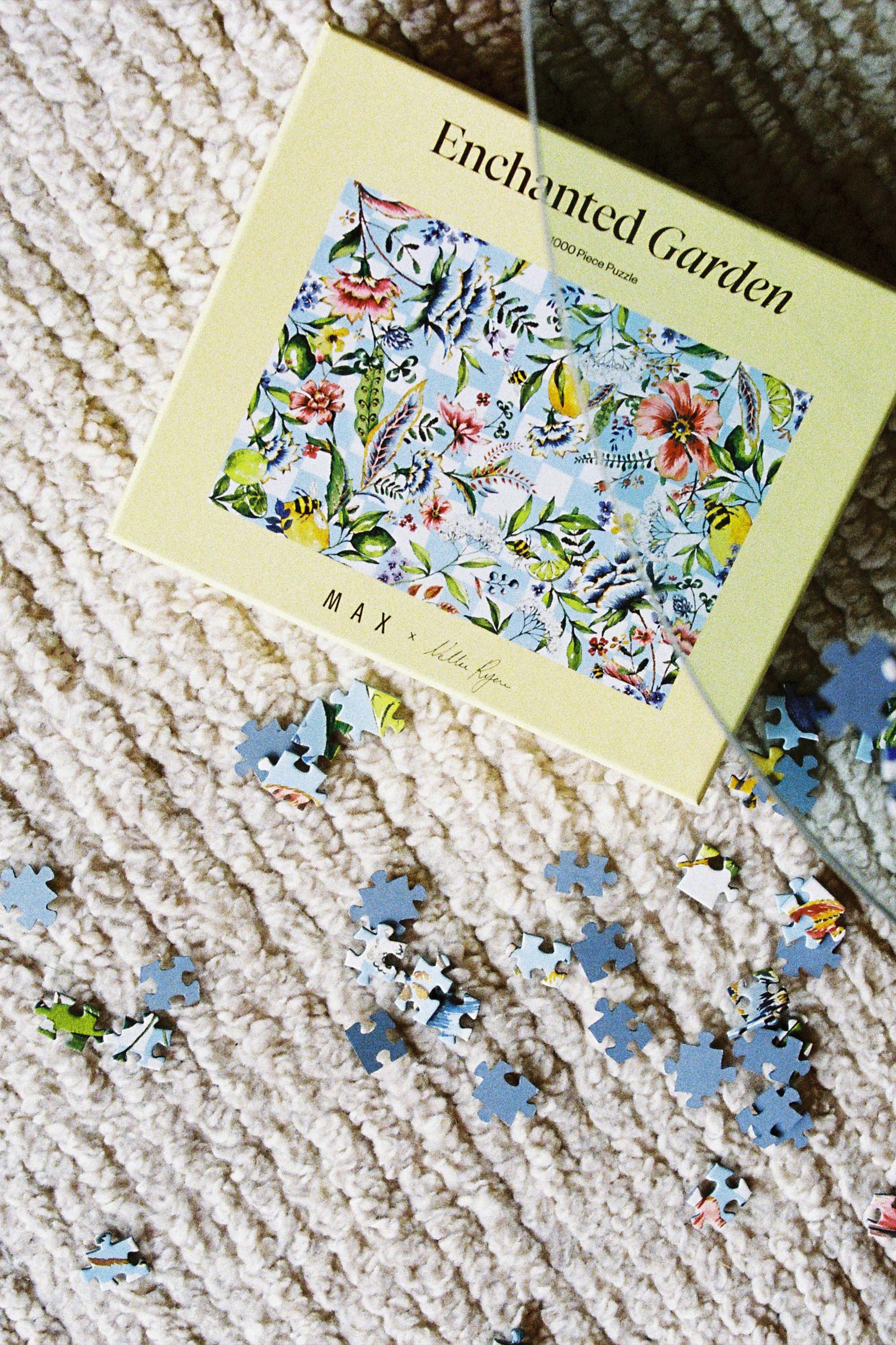 A Piecehouse Enchanted Garden Puzzle box rests on a textured cream rug with scattered pieces around it. The box displays a vibrant floral design, highlighting the exclusive 1000-piece puzzle inside.