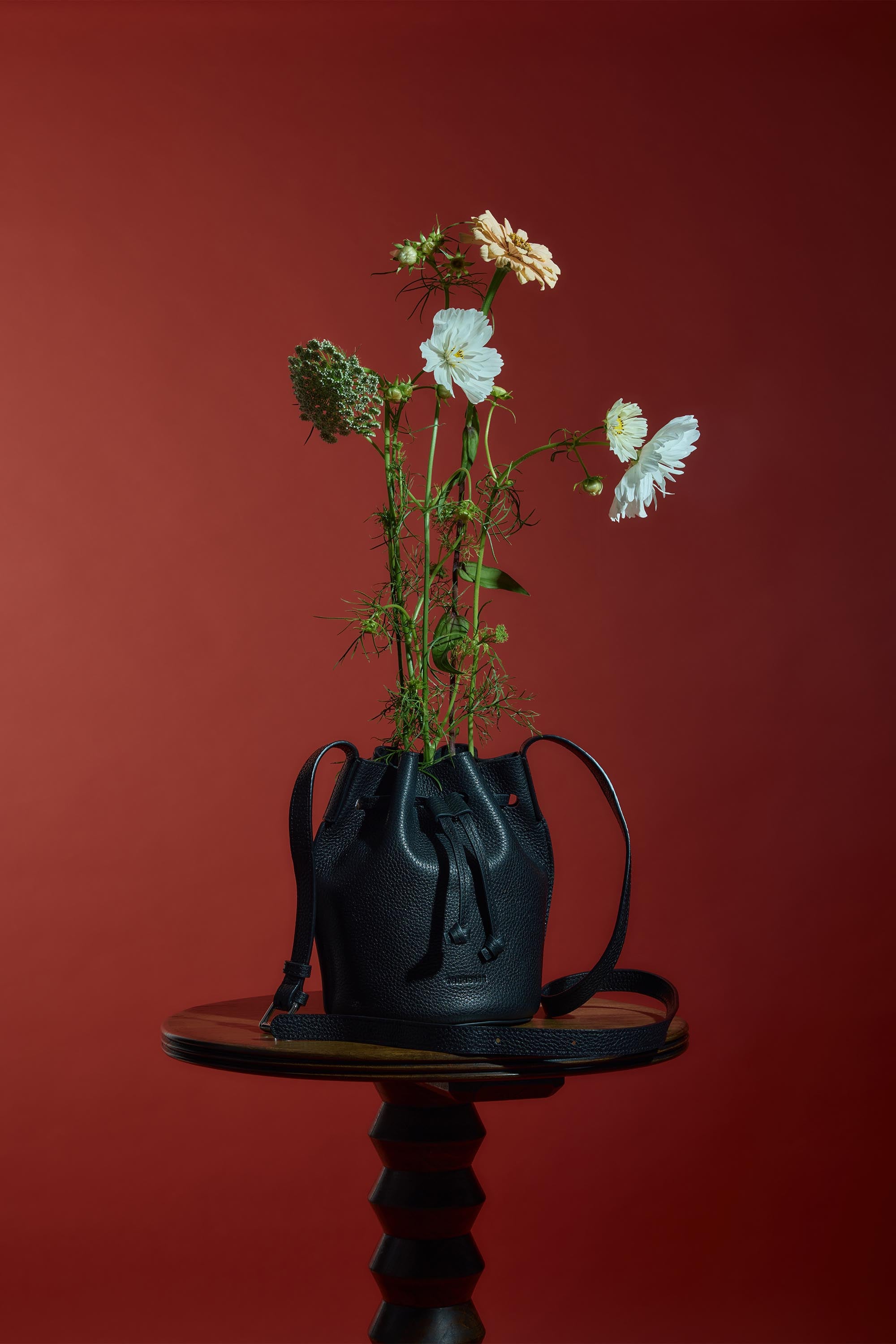 The Horse Bob Bucket, a black grained leather bag by The Horse, is used as a vase for tall flowers on a round wooden table, set against a deep red background.