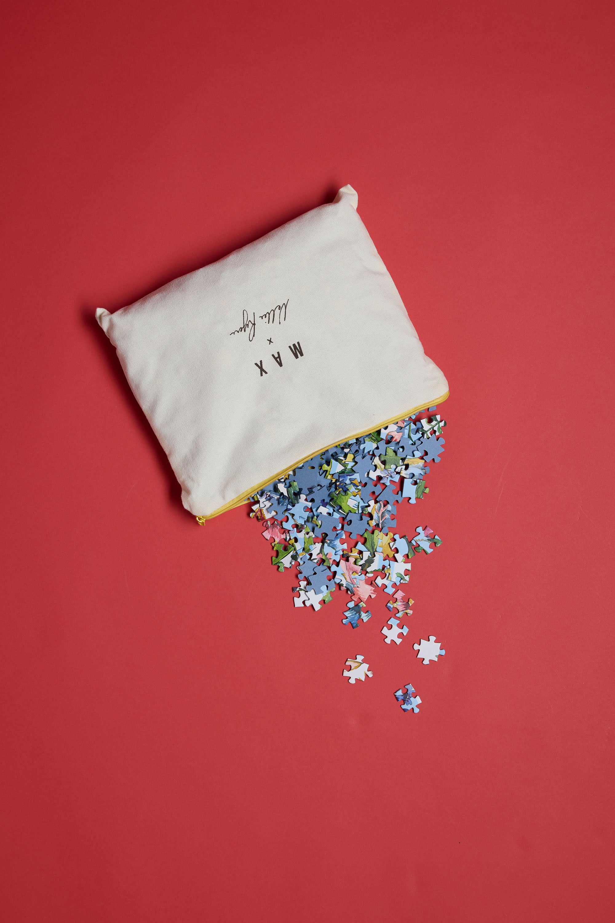 A white Piecehouse pouch with black text sits on a red surface, partially open, with colorful Enchanted Garden Puzzle pieces spilling out—hinting at the exclusive 1000-piece puzzle inside.