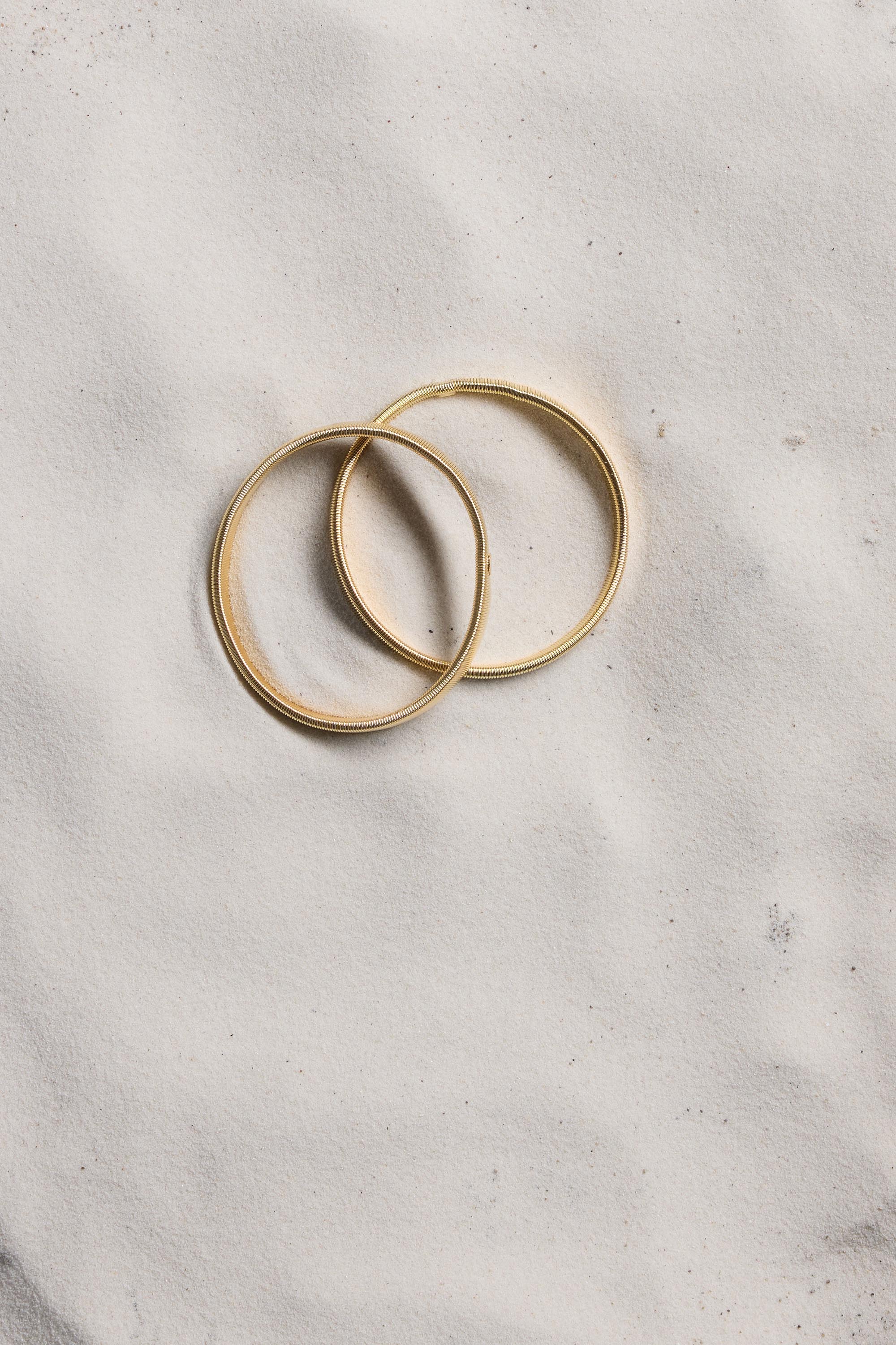Max Sleeve Garter Pair placed on light sand beside two gold wedding rings.