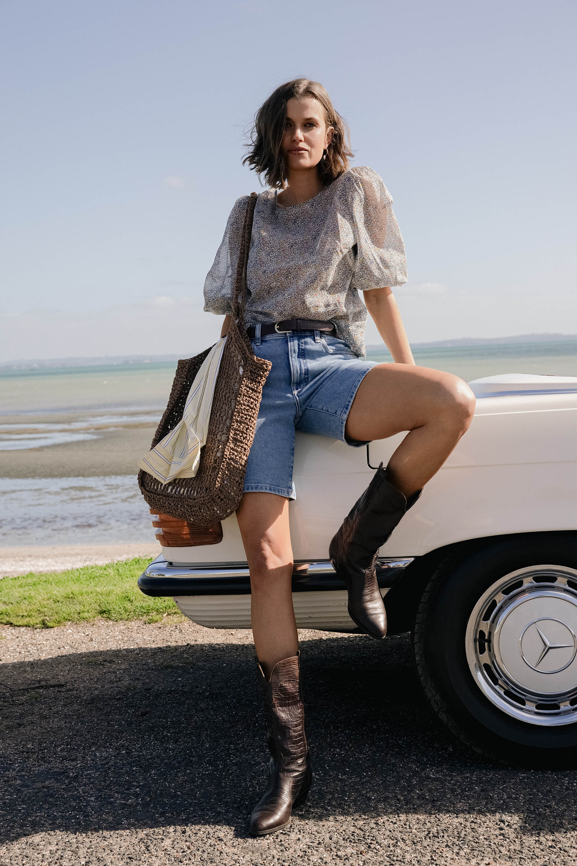 A woman in the Eloise Puff Sleeve Blouse by Max with Liberty Prints floral, denim shorts, and brown cowboy boots leans on a white vintage car, carrying a large woven bag by the seaside on a sunny day.