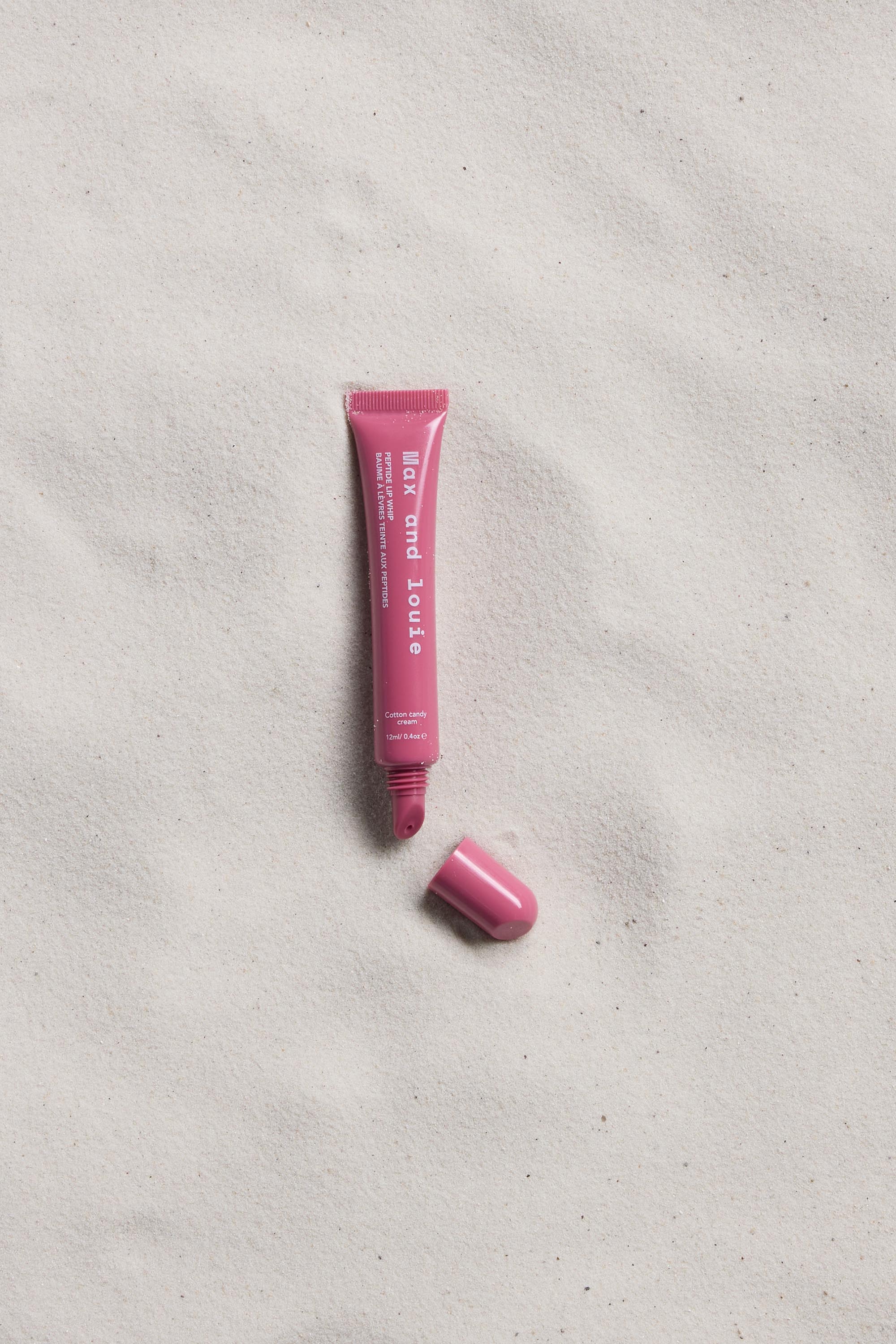 A pink tube of Max and Louie Peptide Lip Whip, with its cap off and resting nearby, is labeled Max and Louie and lies on light, textured sand.