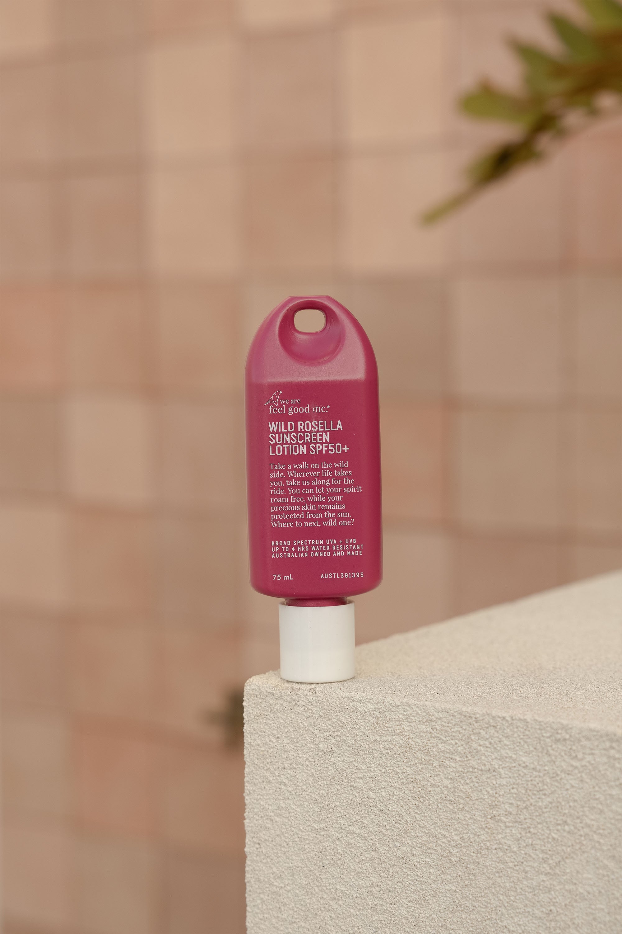 A 75ml pink bottle of We Are Feel Good Signature Sunscreen SPF50+ stands upright on a white textured surface, with a tiled wall and a blurred green plant in the background.