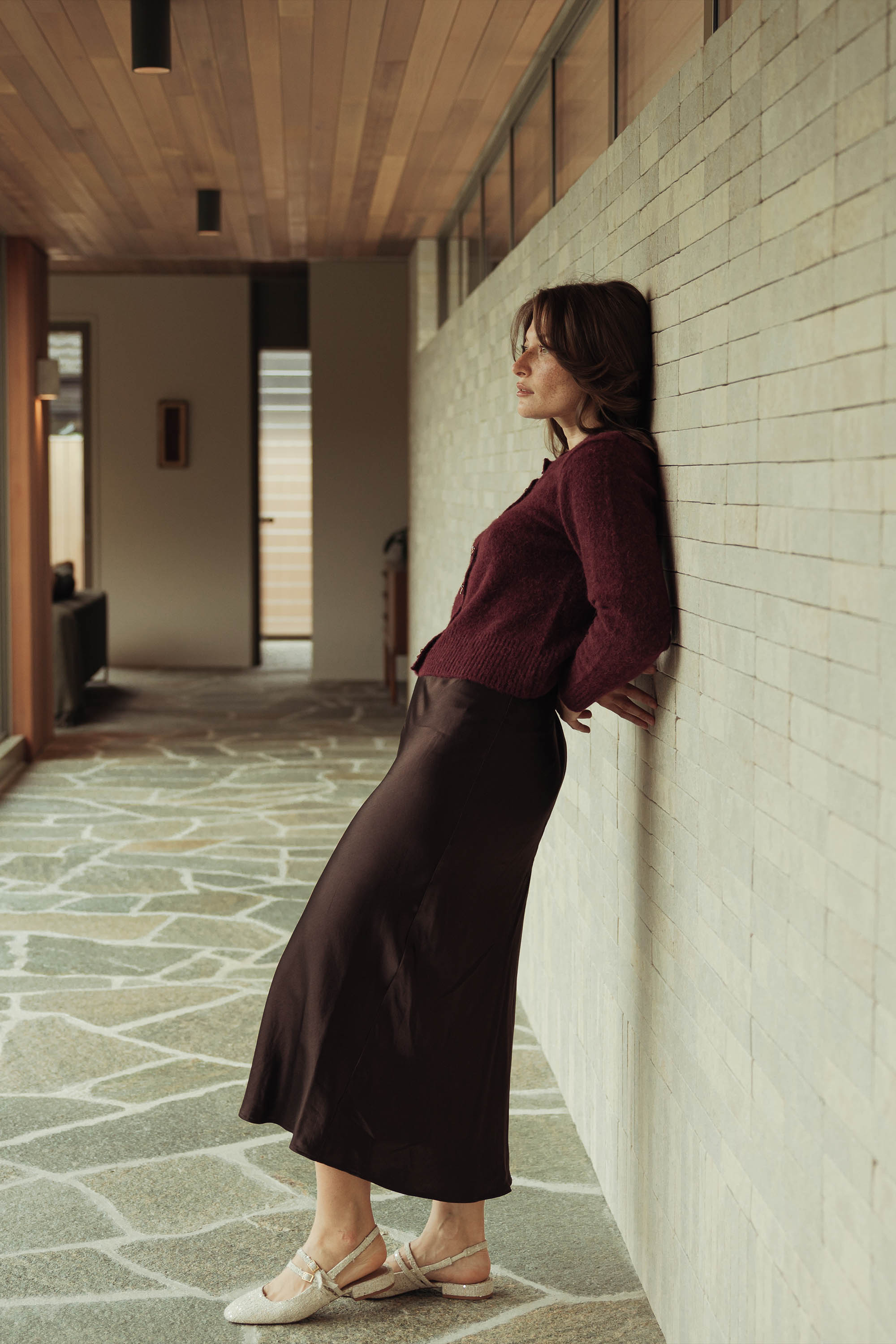A woman in a maroon sweater and Max’s The Slip Skirt leans against a light brick wall in a modern hallway with stone flooring and a wooden ceiling, looking relaxed with her hands behind her back.