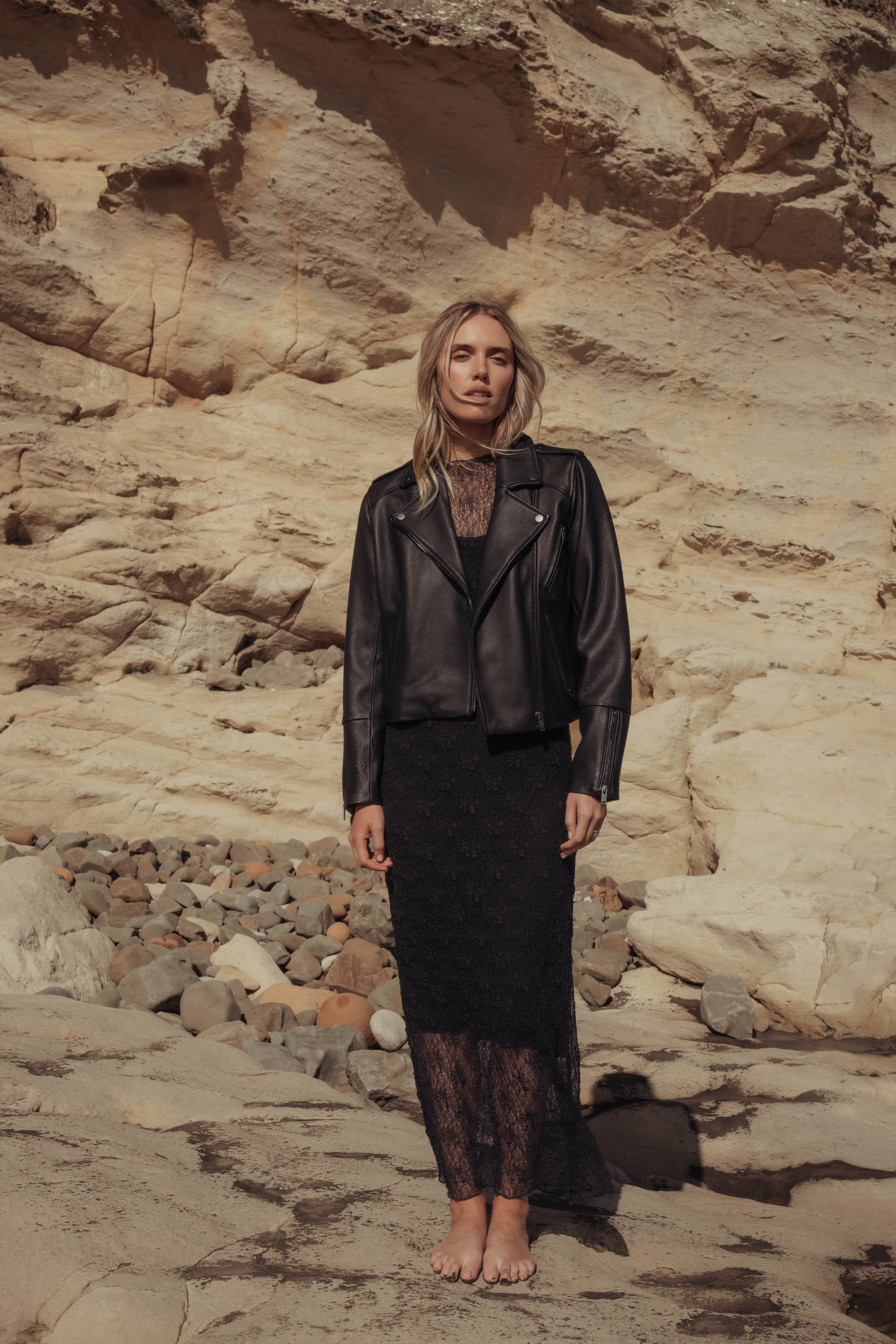 A woman with loose blonde hair and a neutral expression stands barefoot on rocky ground before a beige cliff, wearing the Max "The Perfect Leather Biker Jacket," a timeless staple paired with a black lace dress.