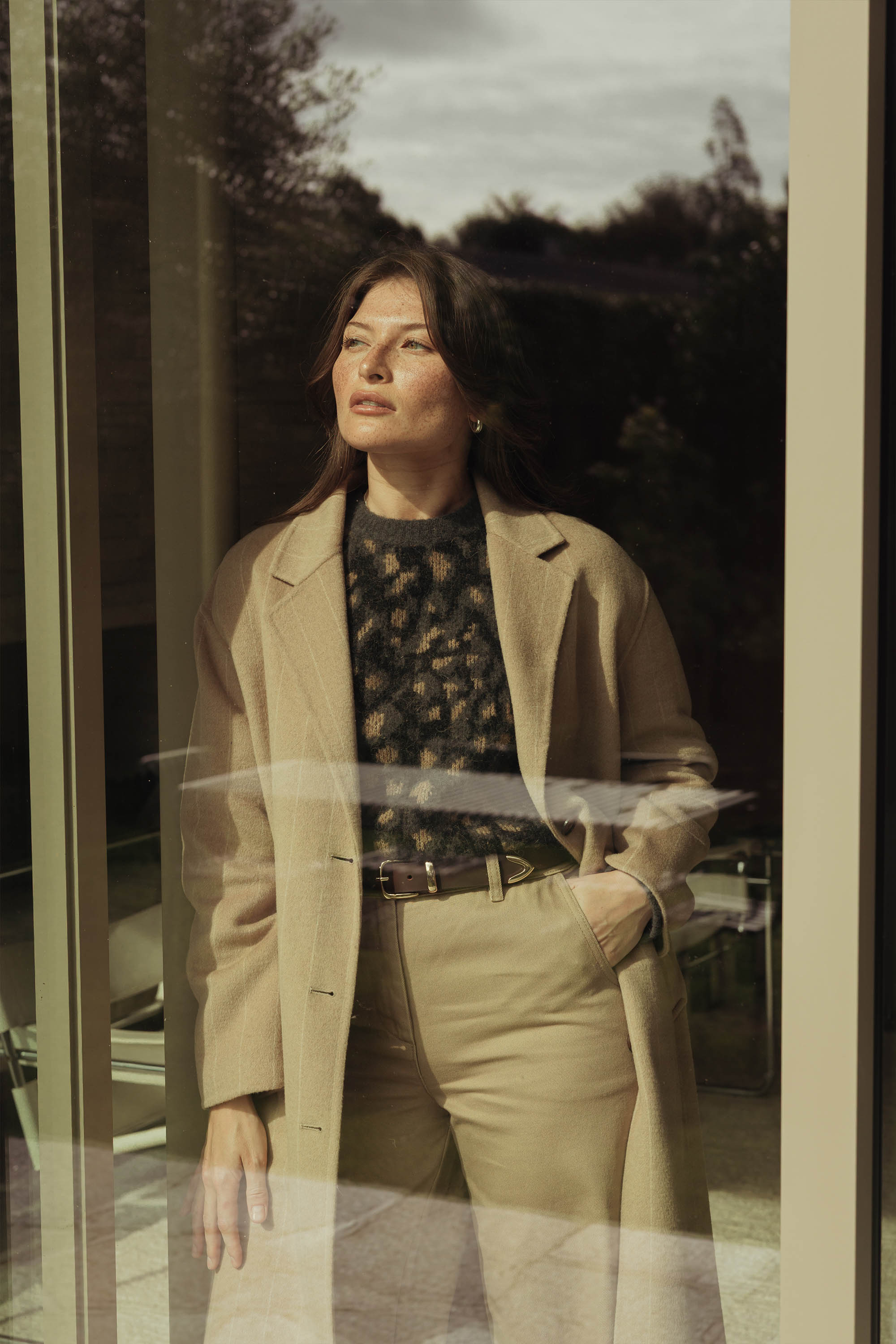 A woman in a beige coat and pants wears the Max Hayley Luxe Whisper Crew Neck Sweater, standing indoors by a window with one hand in her pocket, gazing thoughtfully outside.
