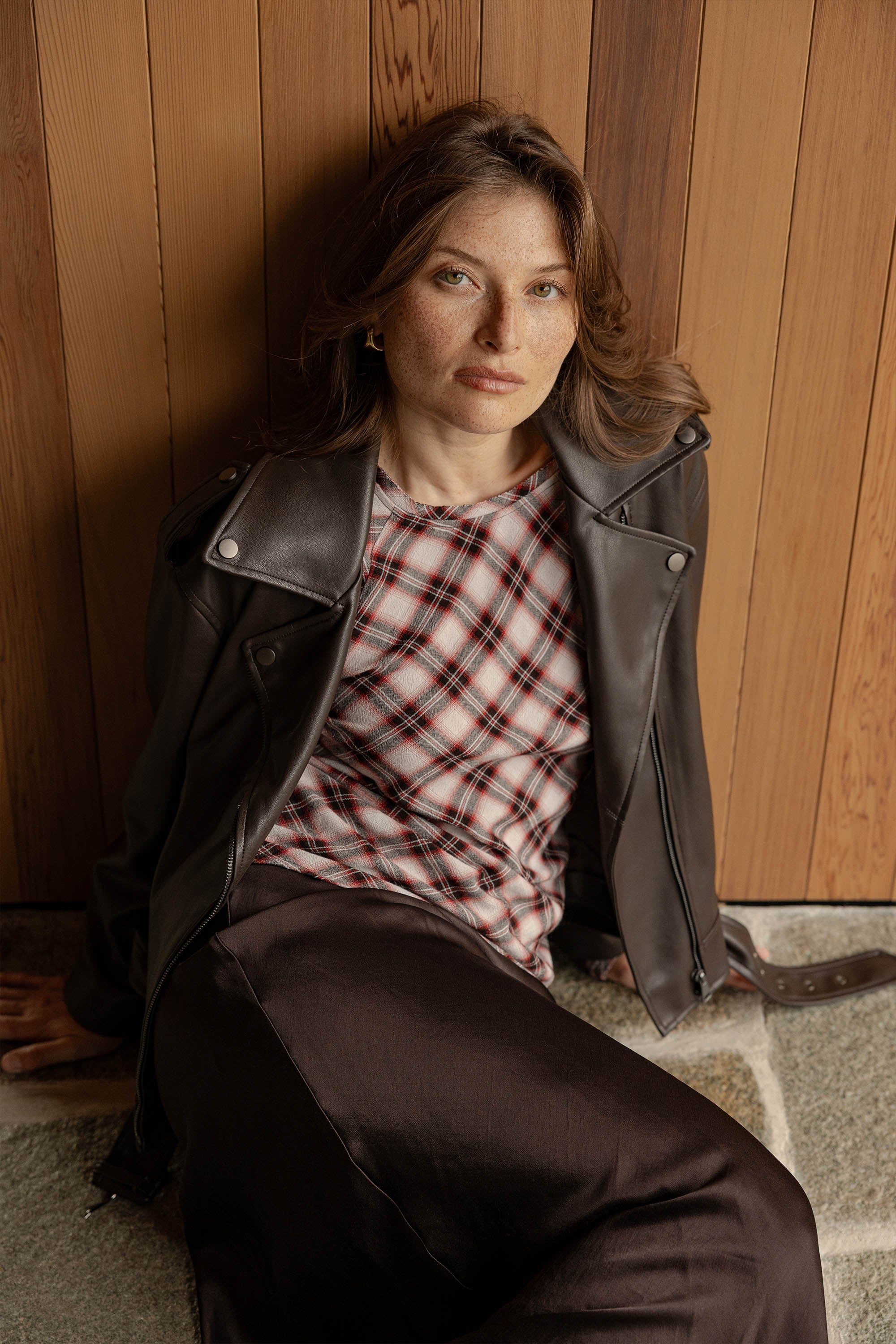 A woman with light brown hair sits against a wooden wall, wearing a black leather jacket over the red Kristen Back Detail Top by Max and a dark skirt, looking calmly at the camera.