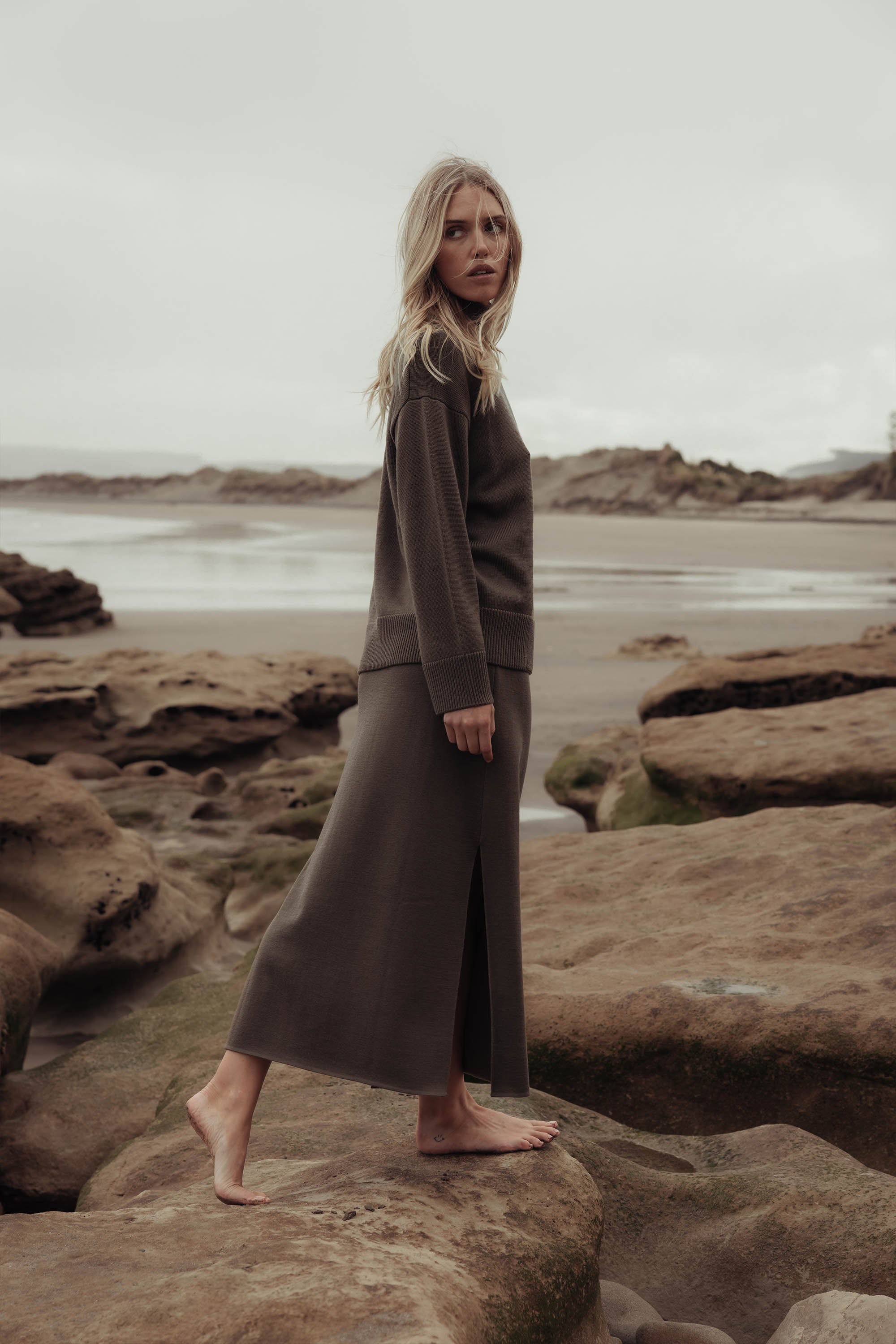 A woman with long blonde hair stands barefoot on rocky shore in an overcast scene, wearing the Elementary Knitted Skirt and matching sweater by Elementary.