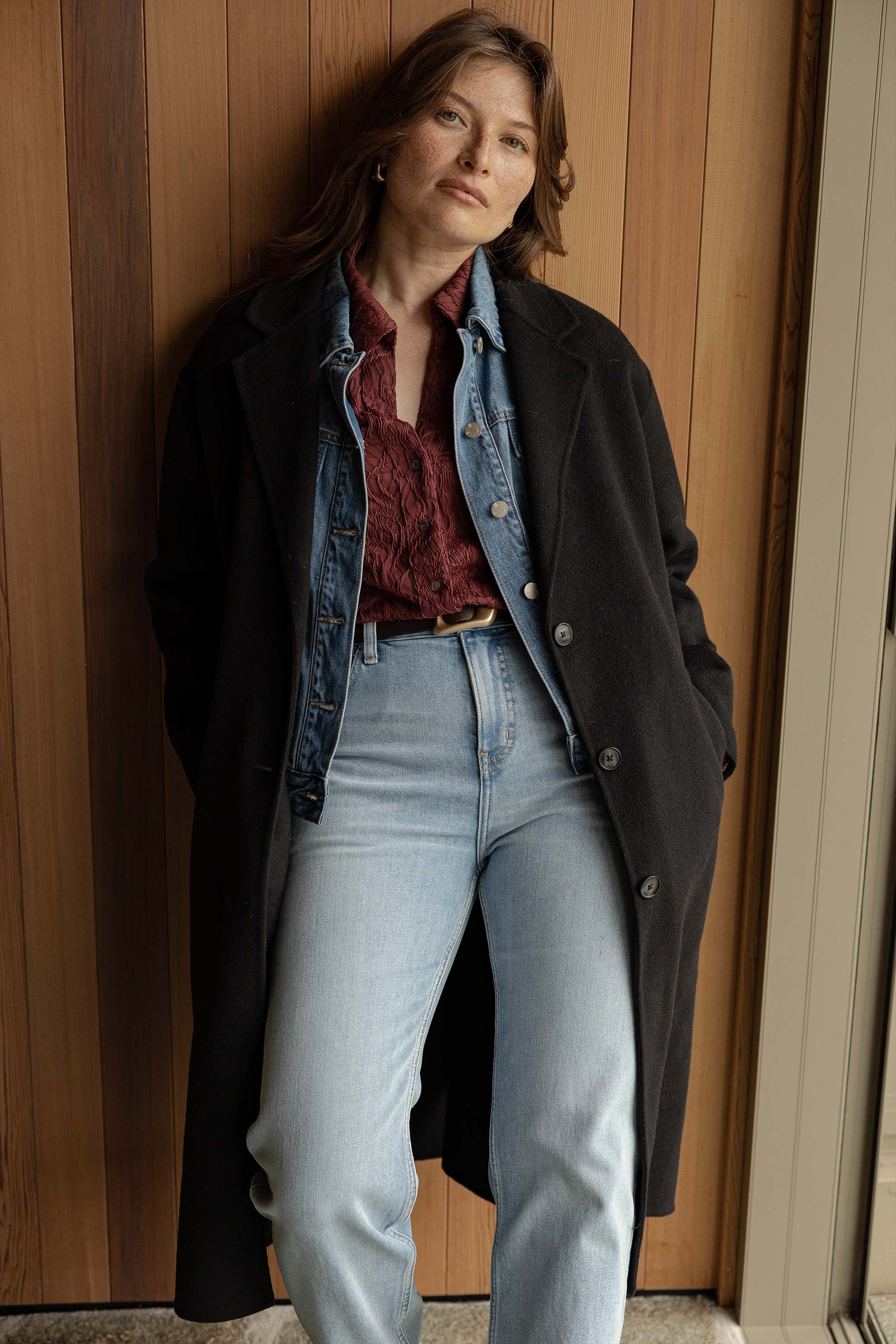 A woman with light brown hair stands by a wooden wall wearing Max's Ella Floral Textured Top under a black coat, paired with a denim jacket and high-waisted light blue jeans. She has a relaxed expression.