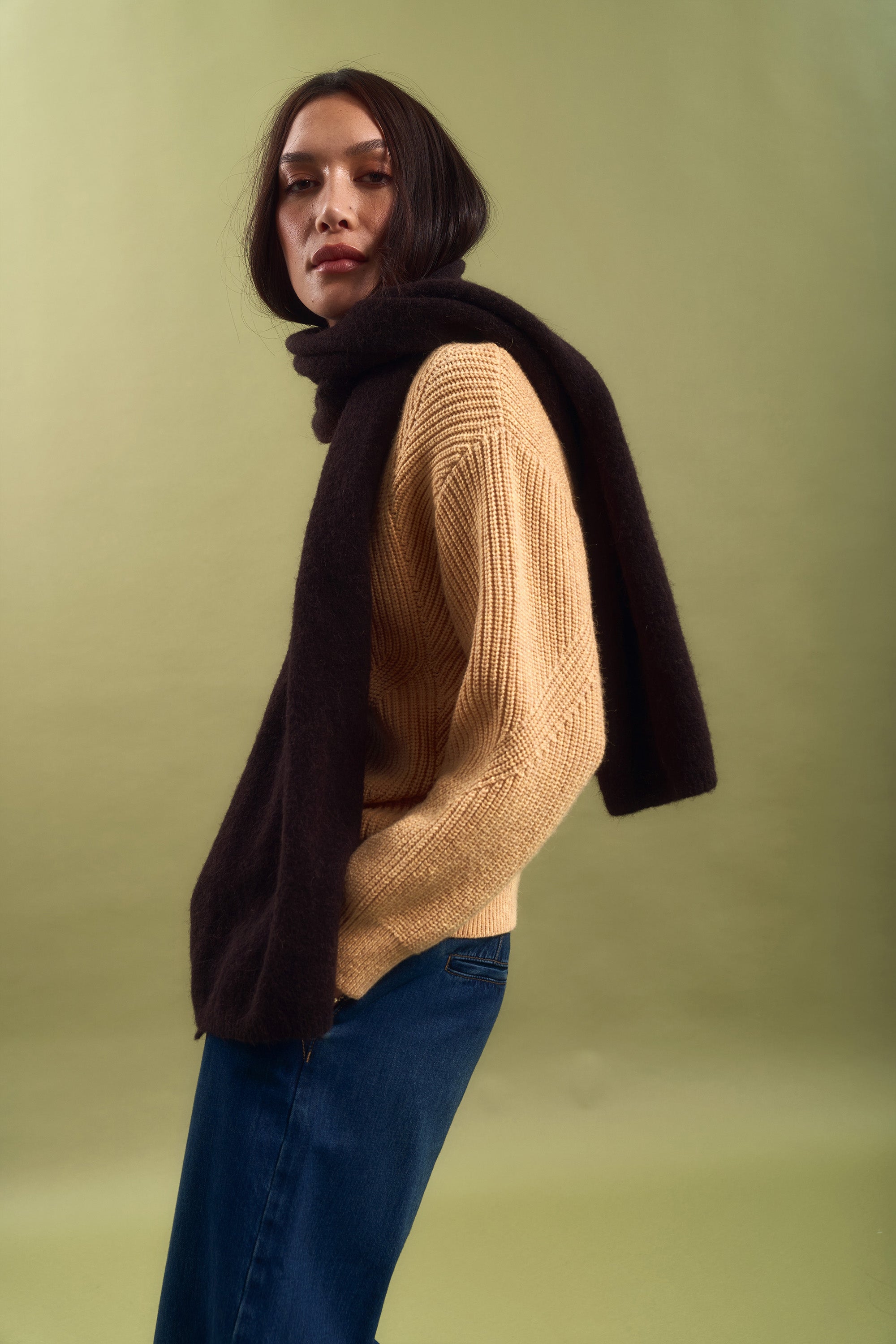 A person wearing the Max Olive Chunky Knit Cardigan stands against an olive-green background, confidently facing the camera, paired with blue jeans and a dark scarf over their shoulders.