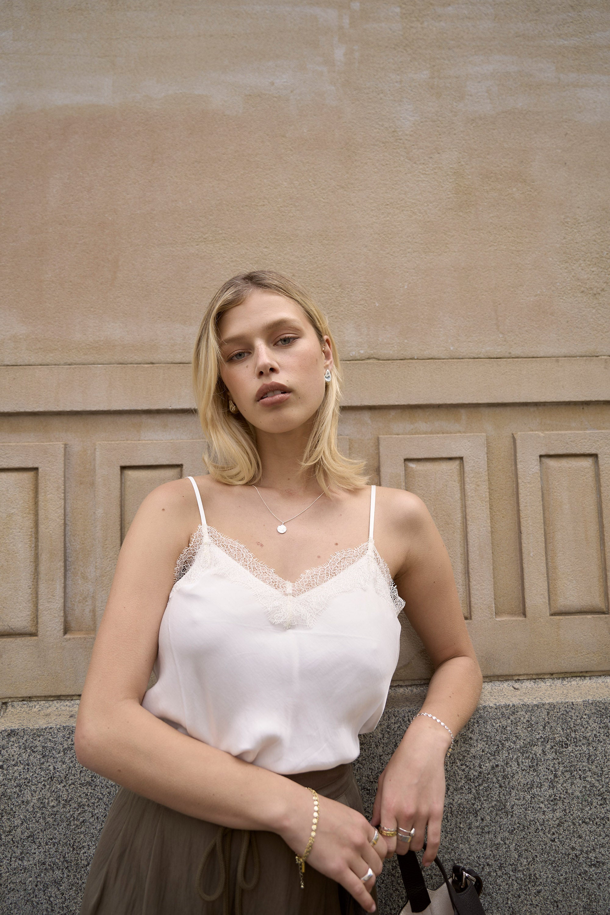 A young woman with blonde hair wears the Max Chloe Lace Detail Tank with adjustable straps and an olive green skirt, standing against a beige textured wall. She gazes confidently at the camera, showcasing minimal jewelry and a neutral expression.