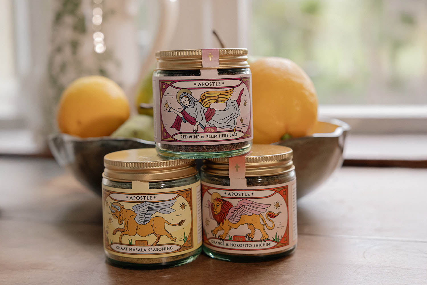 Three small glass jars of Apostle seasoning with illustrated labels are stacked on a table, with a bowl of lemons and limes in the background near a window.