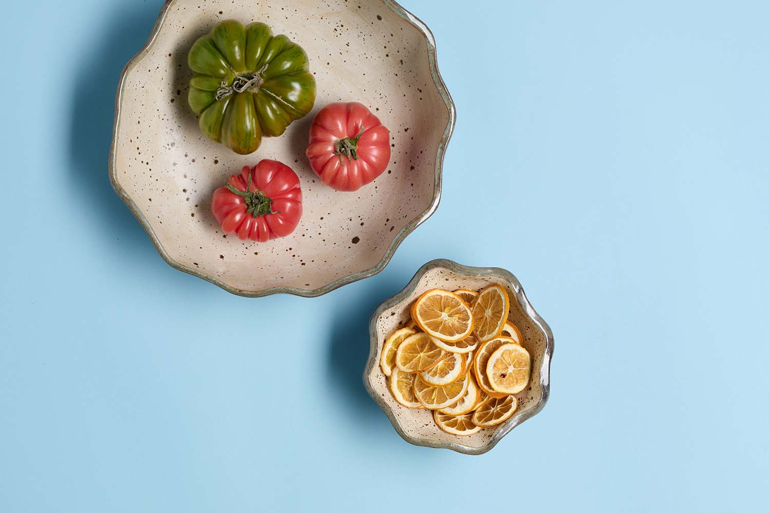 Two ceramic bowls on a blue background; one holds green and red heirloom tomatoes, the other contains dried lemon slices.